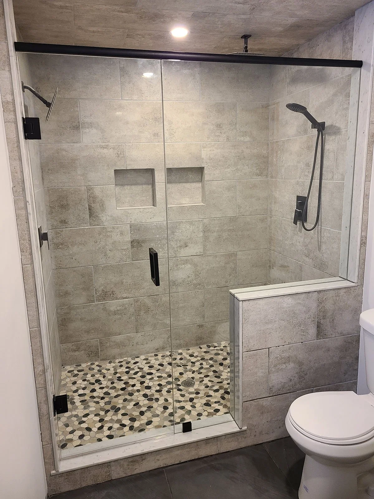 Modern walk-in shower with clear glass doors, beige tile walls, pebble floor, and a handheld showerhead, adjacent to a white toilet.