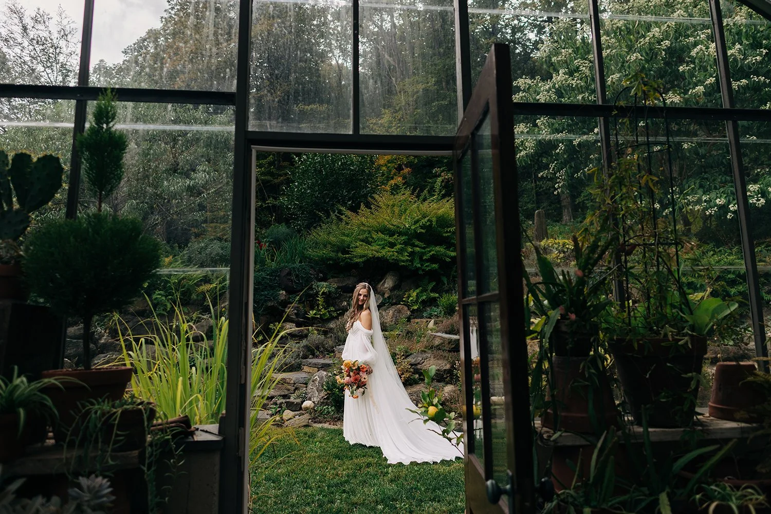 A bride is photographed through the glass panes of a greenhouse during a wedding at western massachusetts wedding venue Gloriosa & Co in Ashfield, Massachusetts.