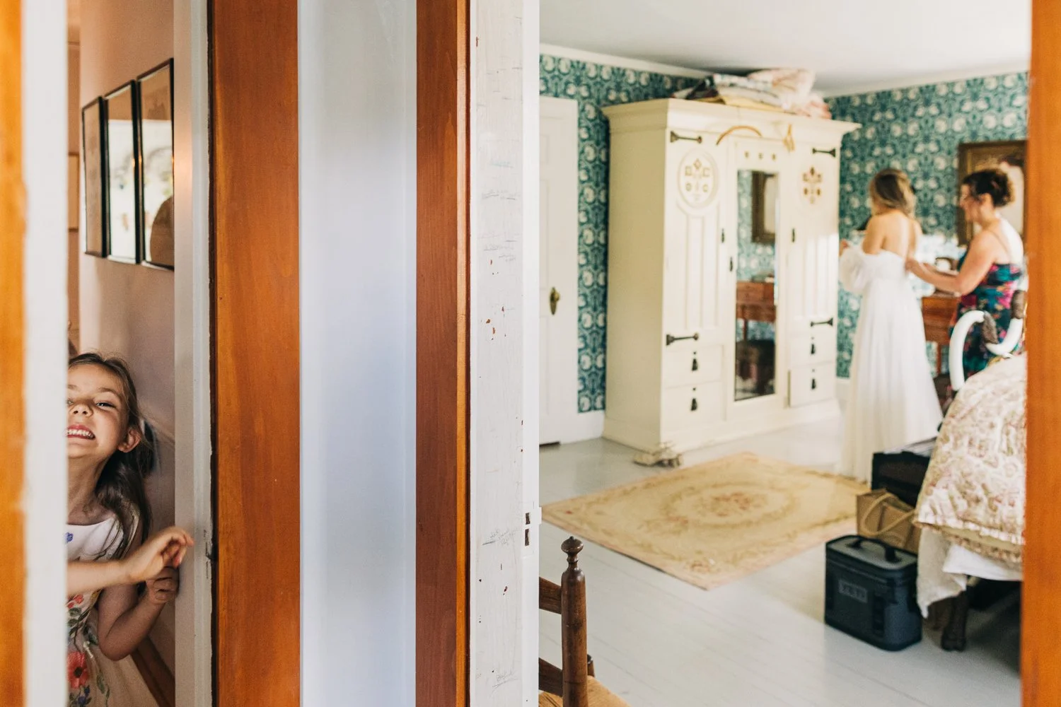 A small child smiles at the camera while peeking through the door to catch a glimpse of the bride during a wedding at western massachusetts wedding venue Gloriosa & Co in Ashfield, Massachusetts.