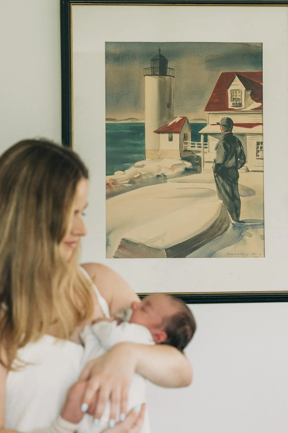 portrait of a mother in front of a painting of a lighthouse during a newborn lifestyle photo session in massachusetts