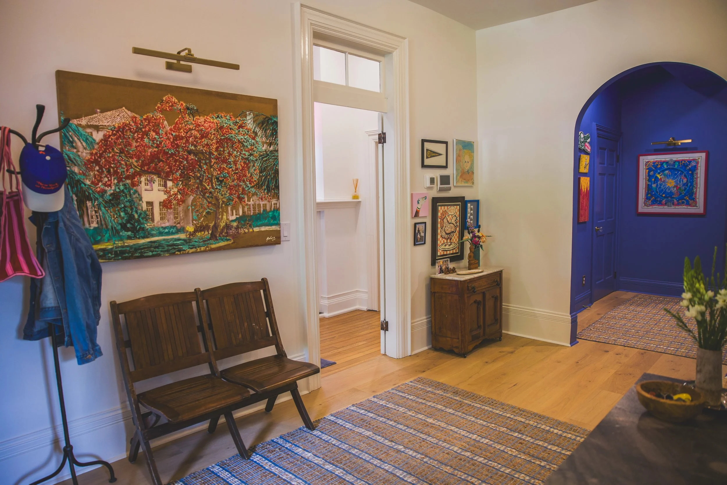 Interior view of a home featuring a hallway with light-colored walls, a wooden bench, colorful artwork on the walls, a small wooden side table with decorations, a blue accent wall in a doorway, a patterned rug, and a large plant in a pot.