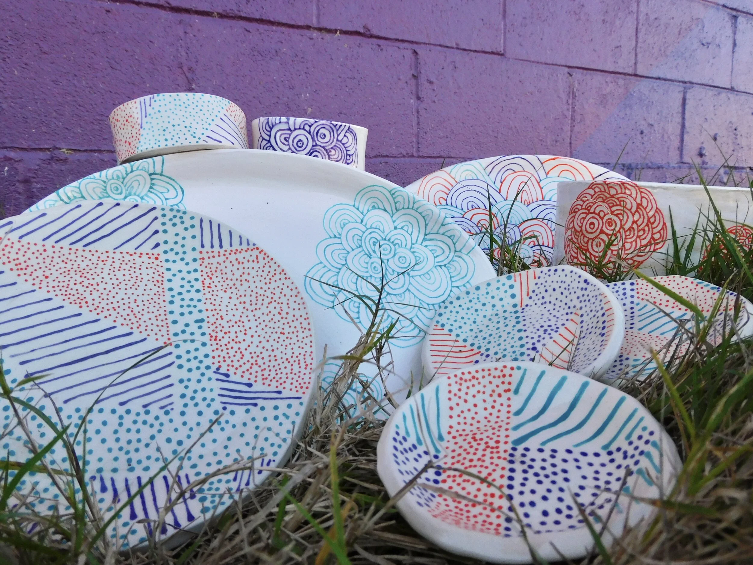 Colorful patterned ceramics placed on the ground among grass in front of a purple brick wall.