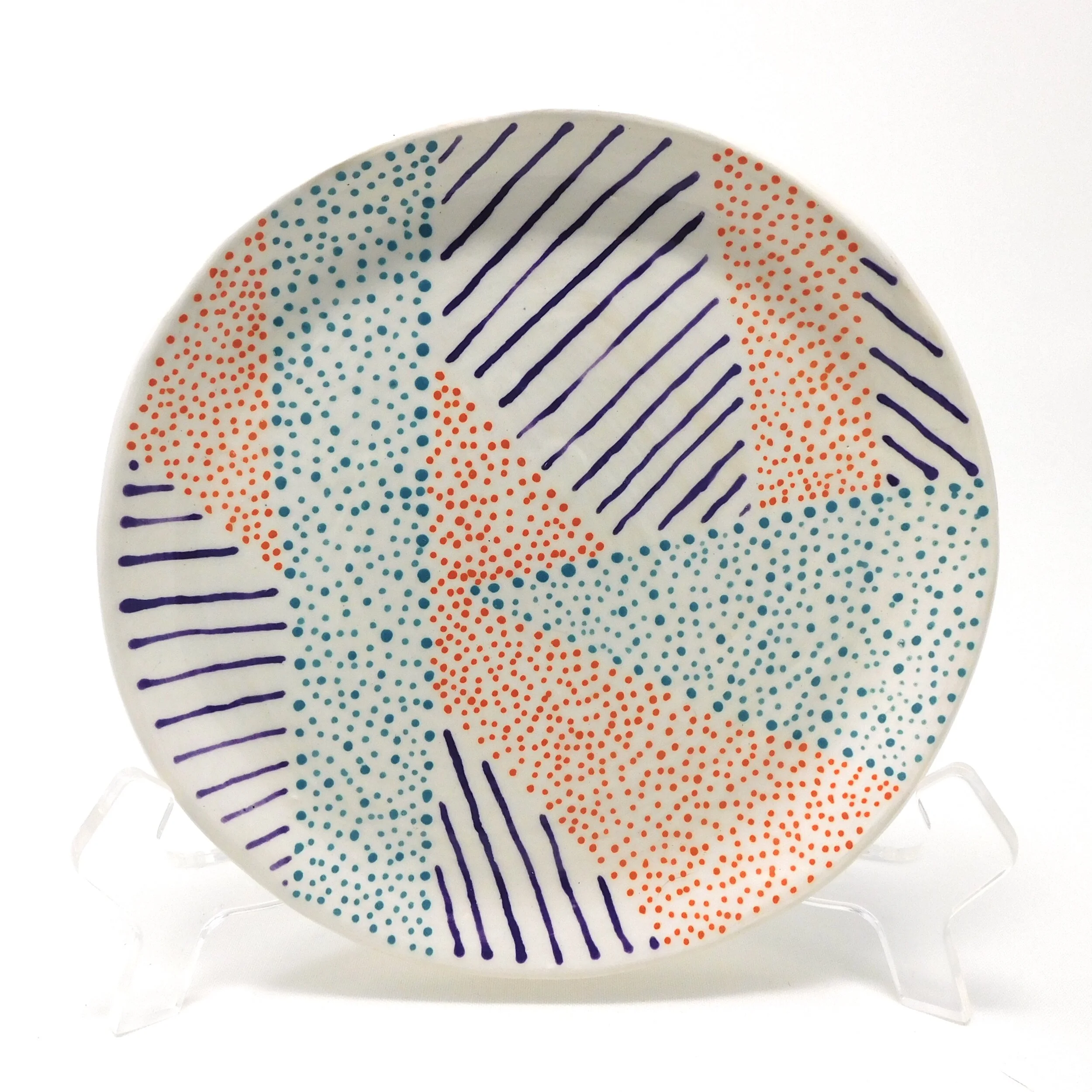 A decorative ceramic plate with abstract patterns in purple, orange, and teal dots and lines on a white background.