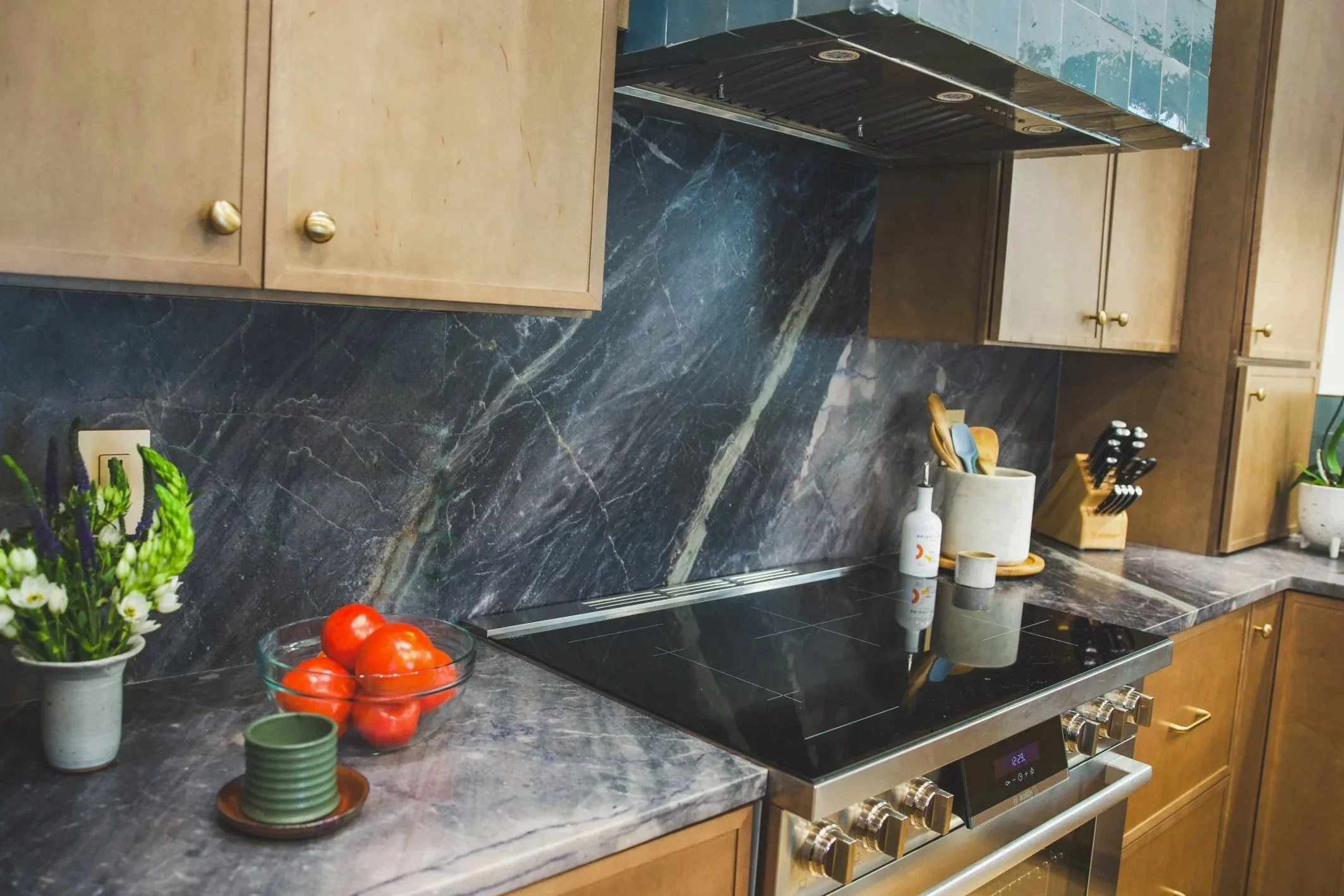 A kitchen countertop with colorful surface, wooden cabinets with brass knobs, a stove with oven controls, a white vase with utensils, a bottle, a potted plant, and a clear glass bowl of tomatoes.