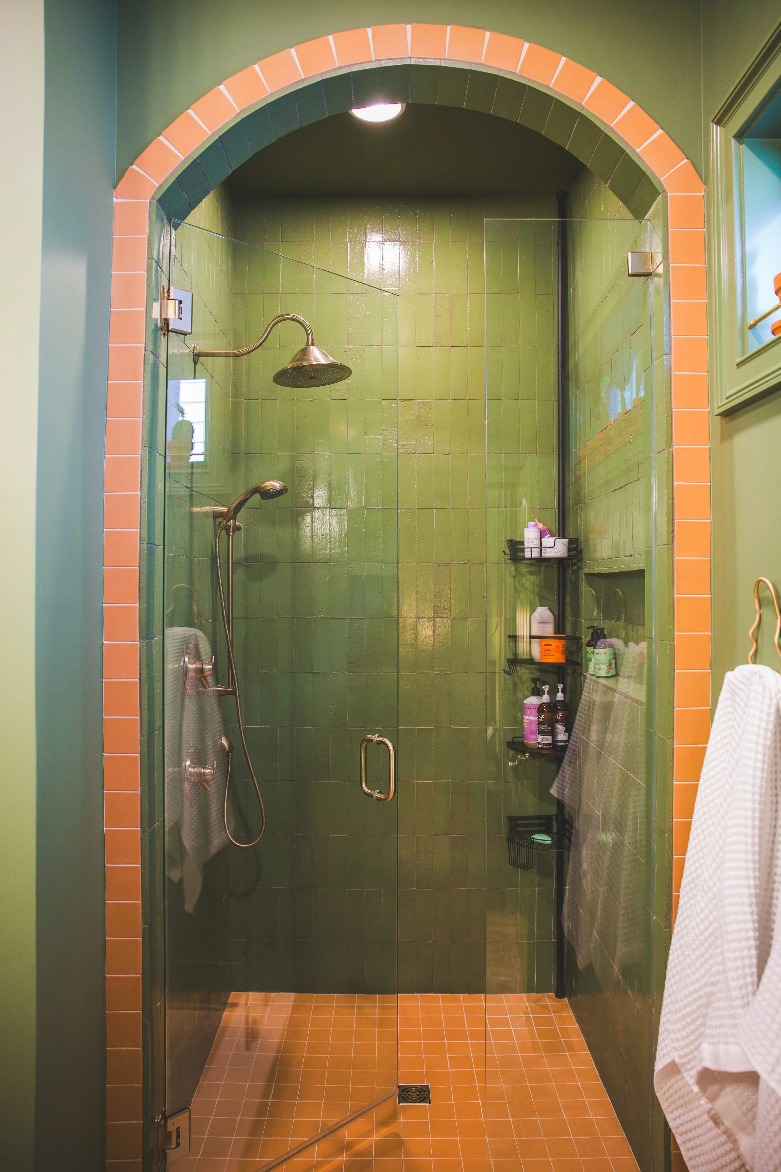 A walk-in shower with green tiled walls, a rain showerhead, a handheld showerhead, and a black metal shelf holding toiletries. The shower is framed by an orange-tiled arch, with a green and light green wall surrounding it. A towel hangs on the right 