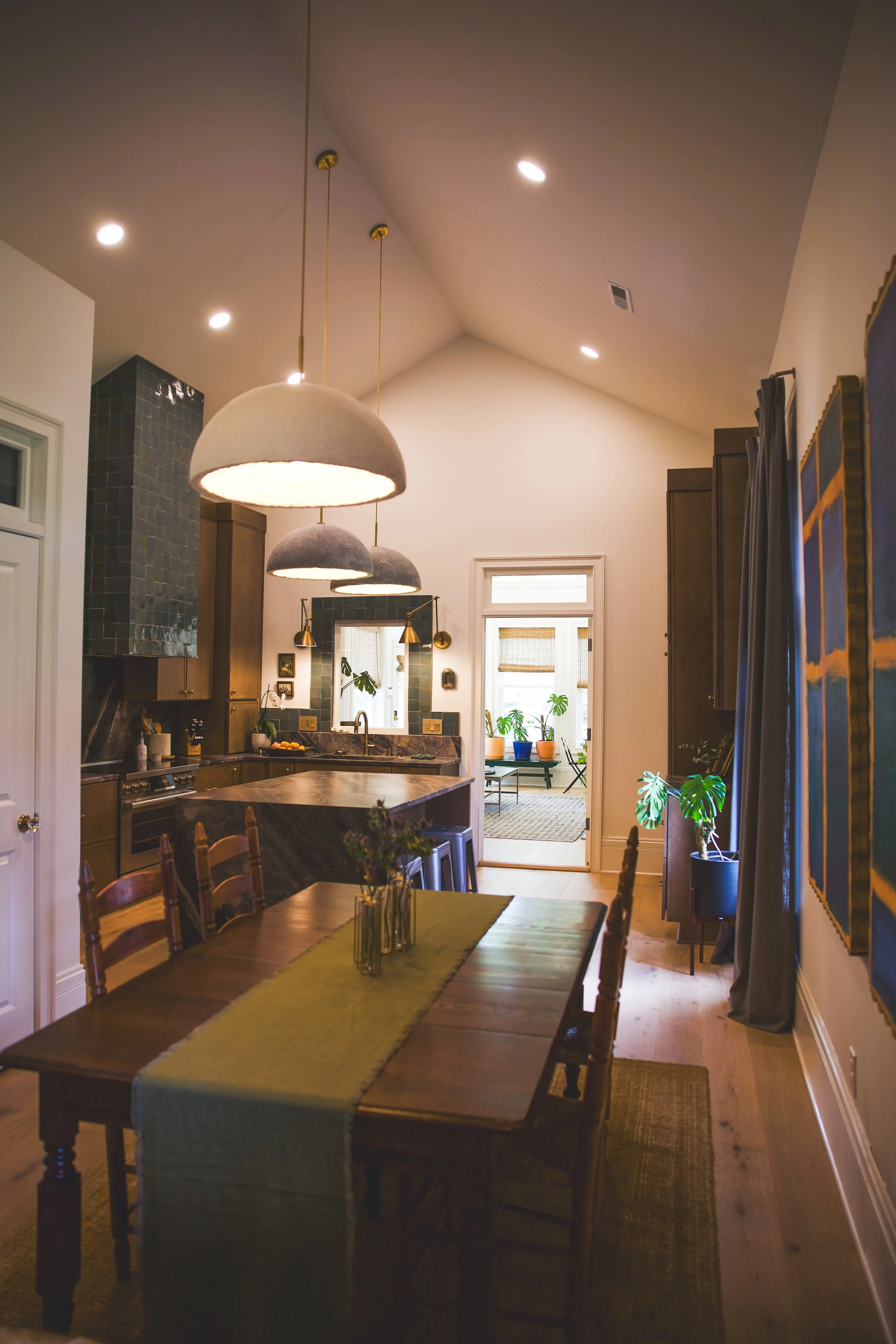The image shows a dining area with a large wooden table decorated with a green table runner, surrounded by wooden chairs. In the background, there is a kitchen with dark cabinetry, a marble countertop, pendant lights hanging from the ceiling, and a w