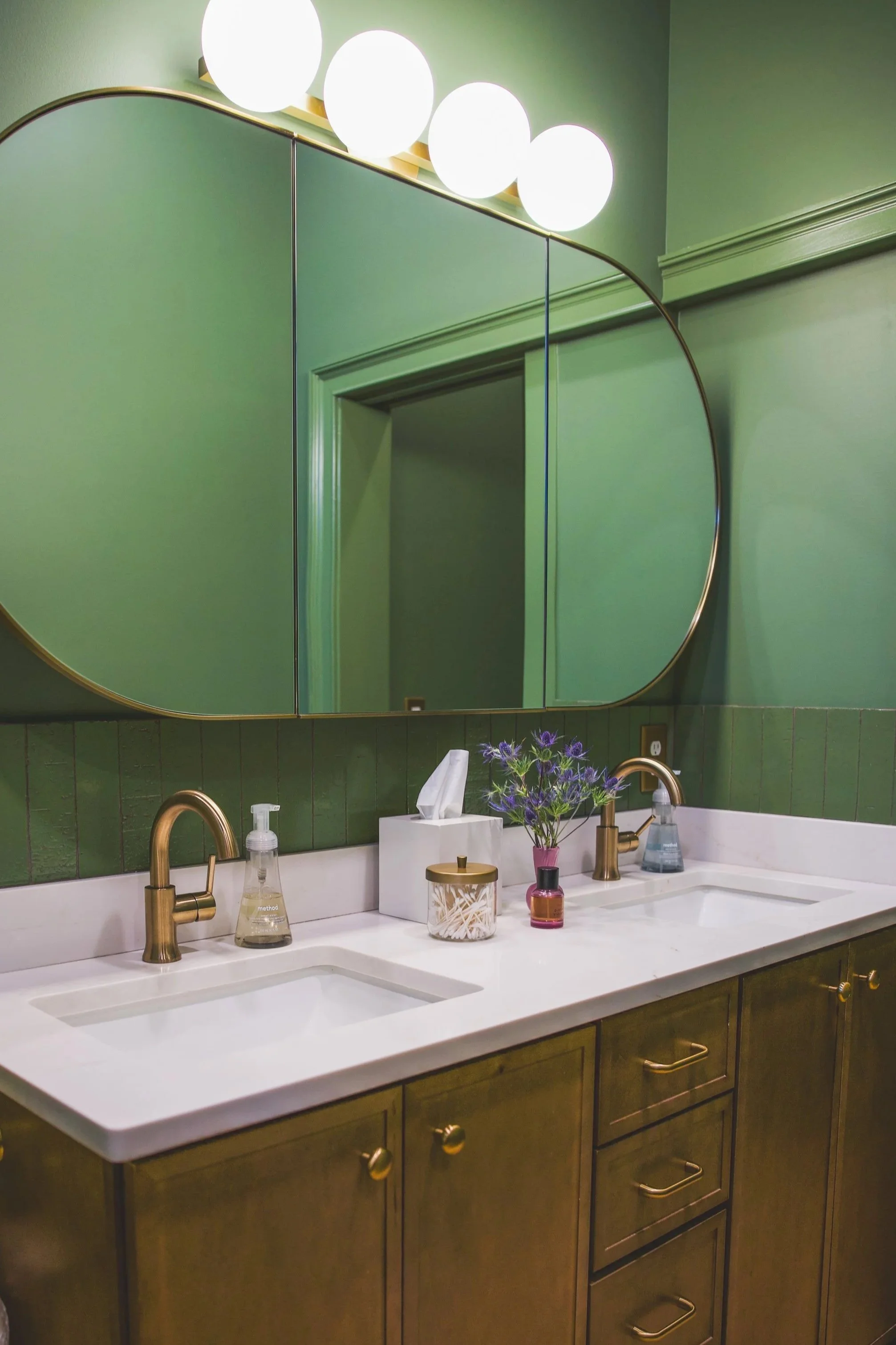 A bathroom vanity with two sinks, brass fixtures, a large mirror, green walls, and decorative items including a vase with purple flowers, a tissue box, and various soap dispensers.