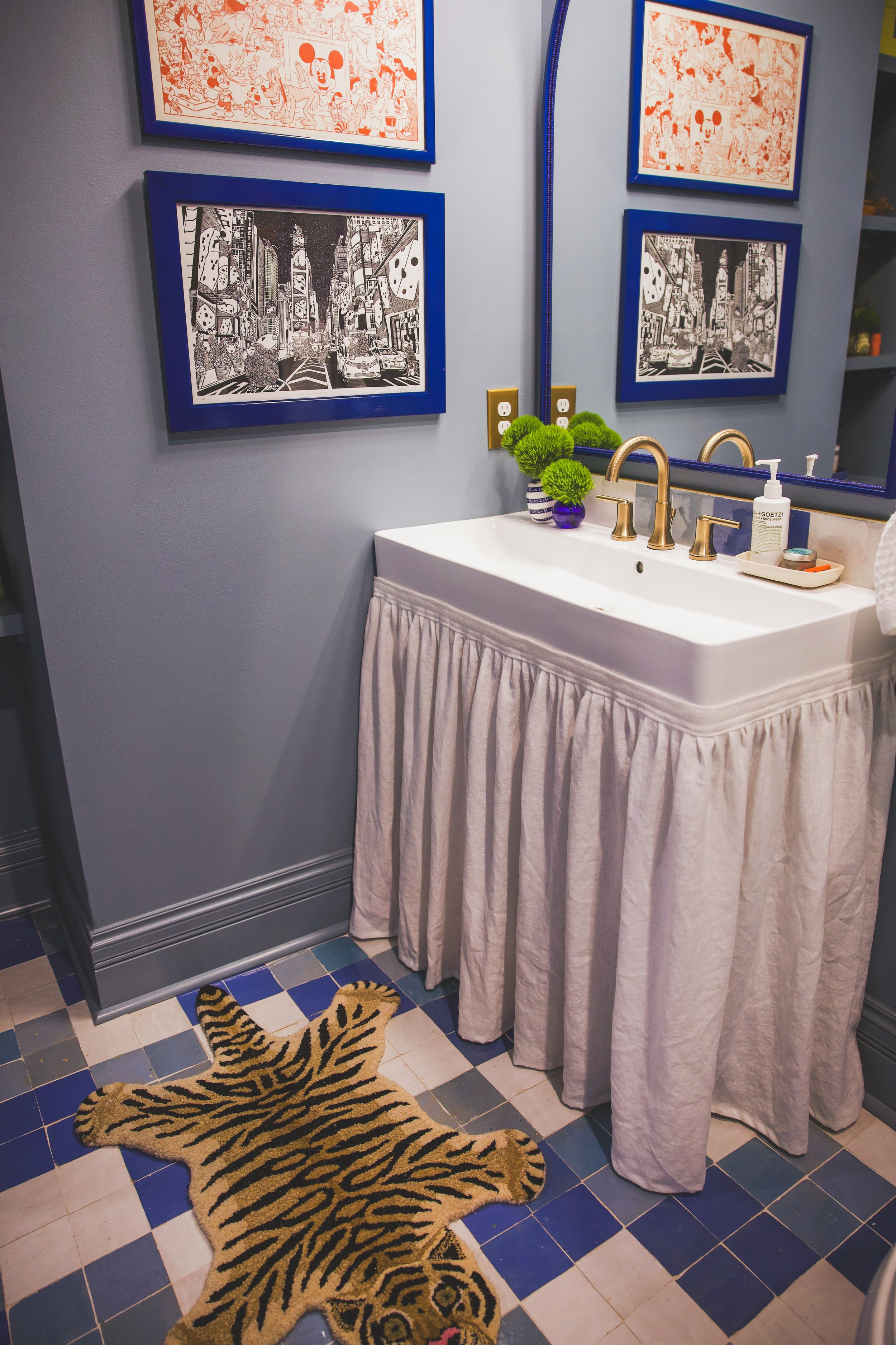 A bathroom with a vanity sink, faucet, two potted green plants, a soap dispenser, and a mirror. Four framed pictures hang on the wall, depicting various art styles including Mickey Mouse and cityscapes. A tiger-shaped rug is on the blue and white che