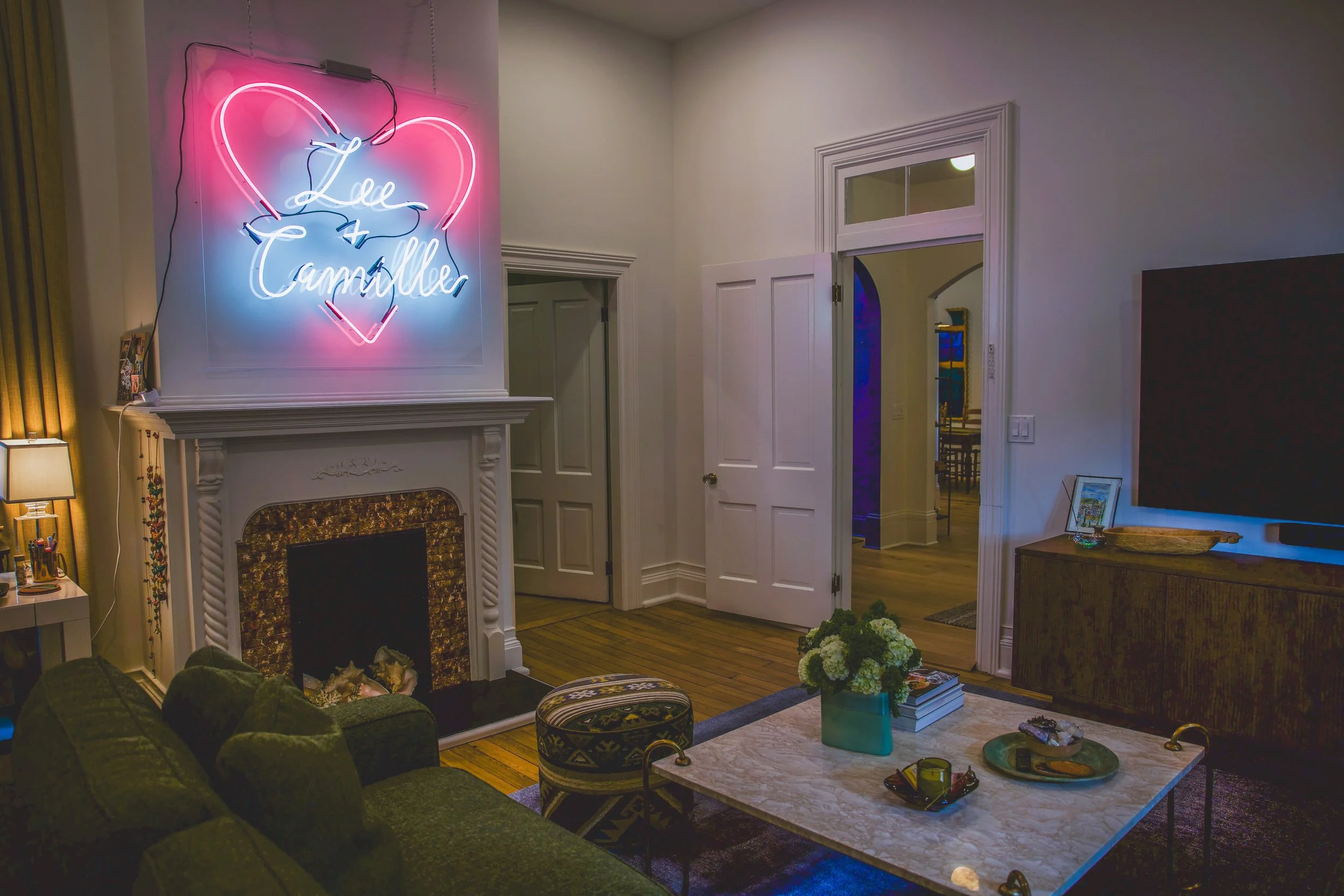 Living room with a fireplace, green velvet sofa, and a coffee table with decorative items. Neon sign on the wall reads 'Lee + Camille' inside a pink heart. There are framed pictures on a side table and decorative flowers on the coffee table.