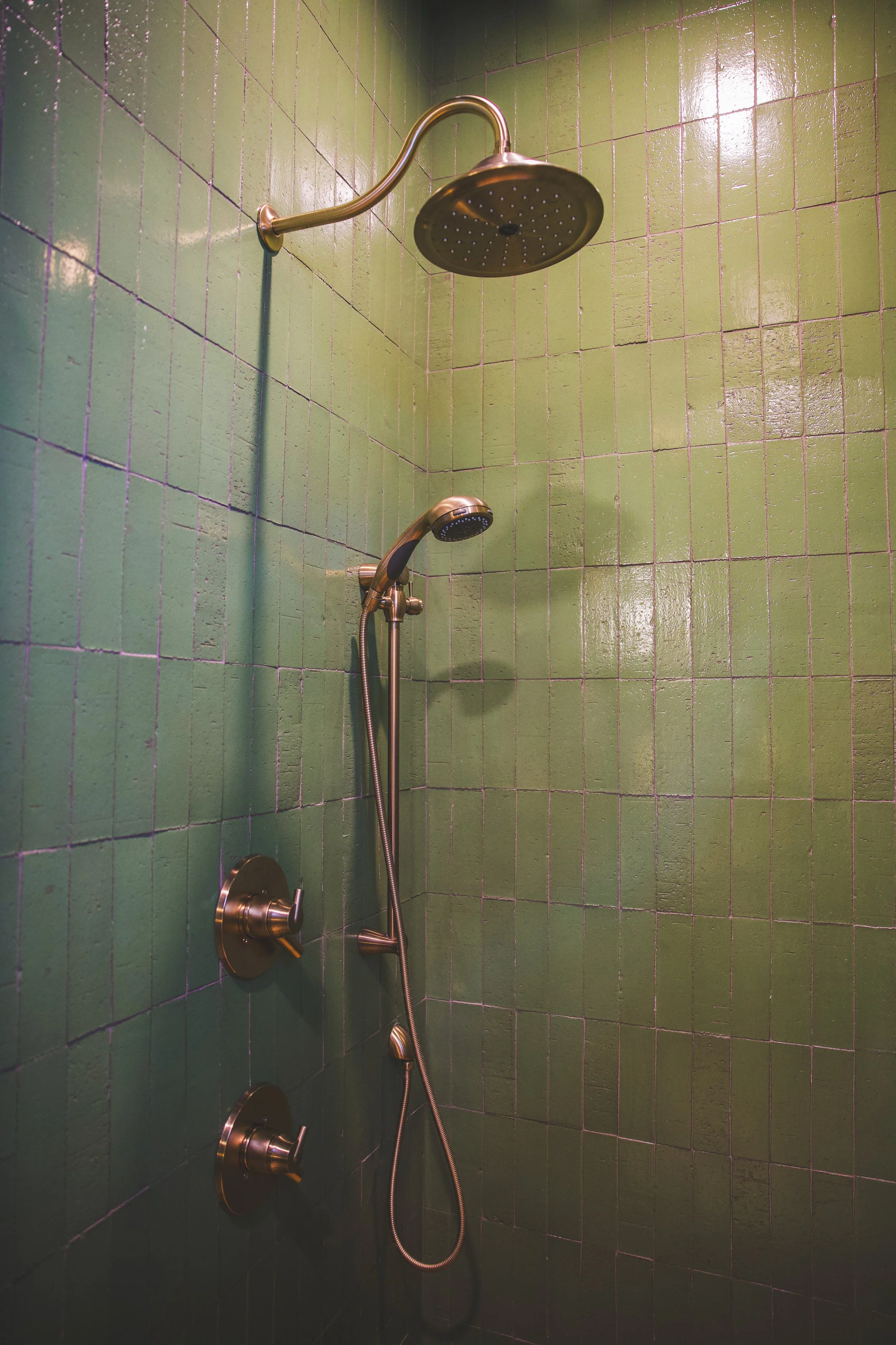 A shower with green tiled walls, a large overhead showerhead, and a handheld showerhead attached by a flexible hose.