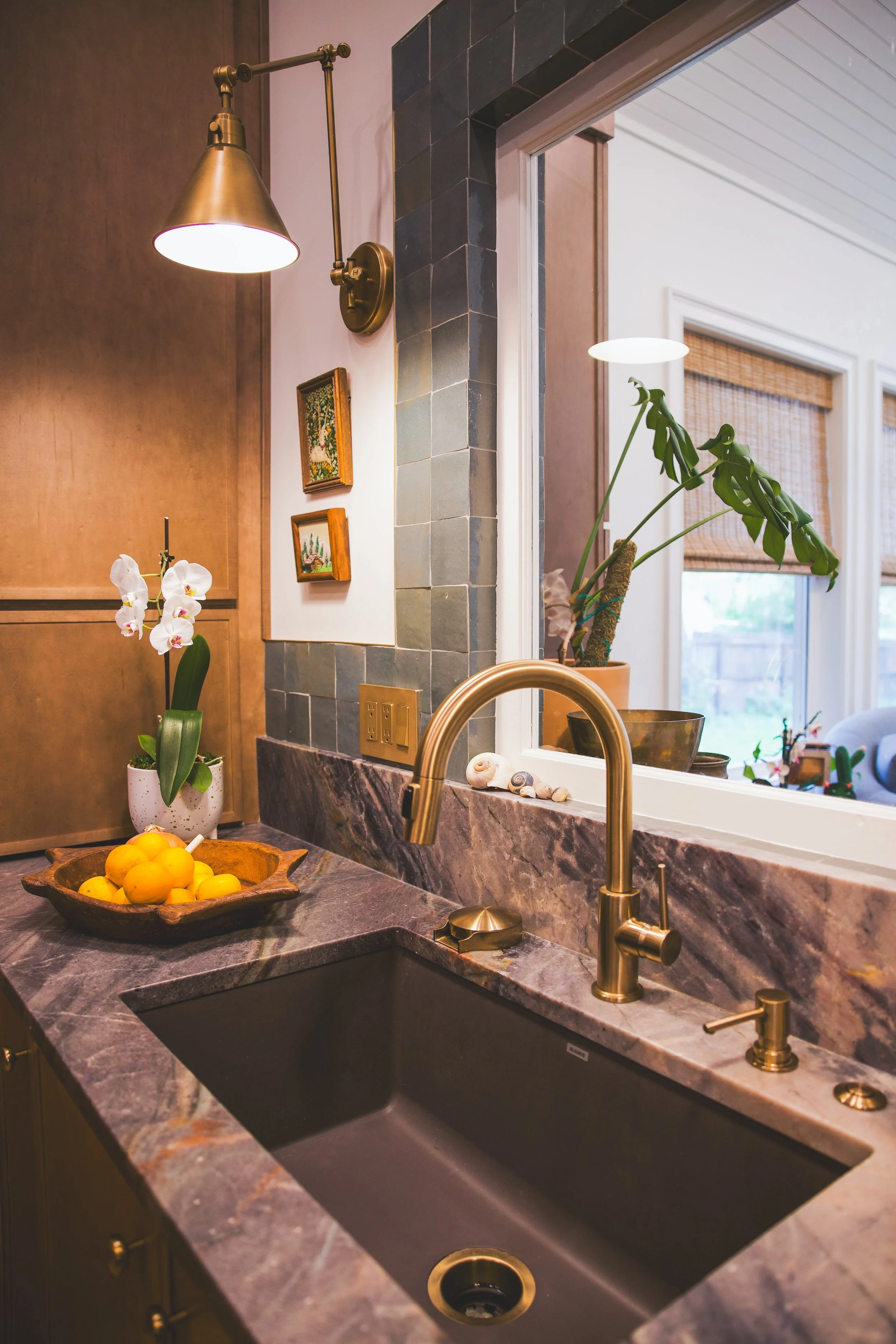 A kitchen countertop with a dark undermount sink, faucet, and a bowl of lemons. A potted orchid and seashells decorate the counter. In the background, a wall with two small framed pictures and a large window leads to a bright room with window shades.