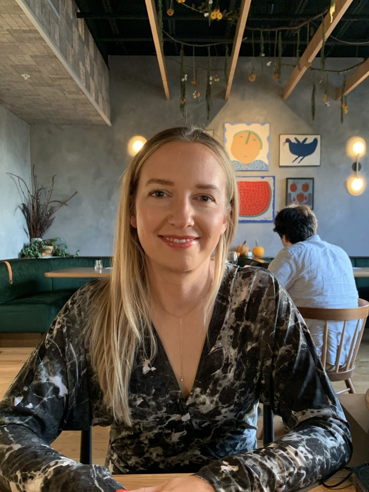 A woman with long blonde hair smiling at the camera in a restaurant. She is wearing a black and white patterned top. The background features colorful abstract artwork on the wall, a man with dark hair sitting at a table with pumpkins, and warm lighting.
