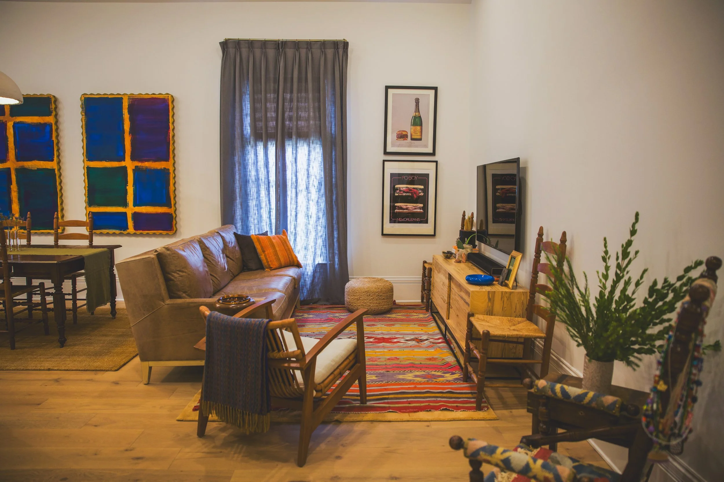 Living room with beige sofa, colorful pillows, wooden chairs, abstract paintings, gray curtains, framed posters, TV on wooden console, patterned multicolored rug, and various decorative items.