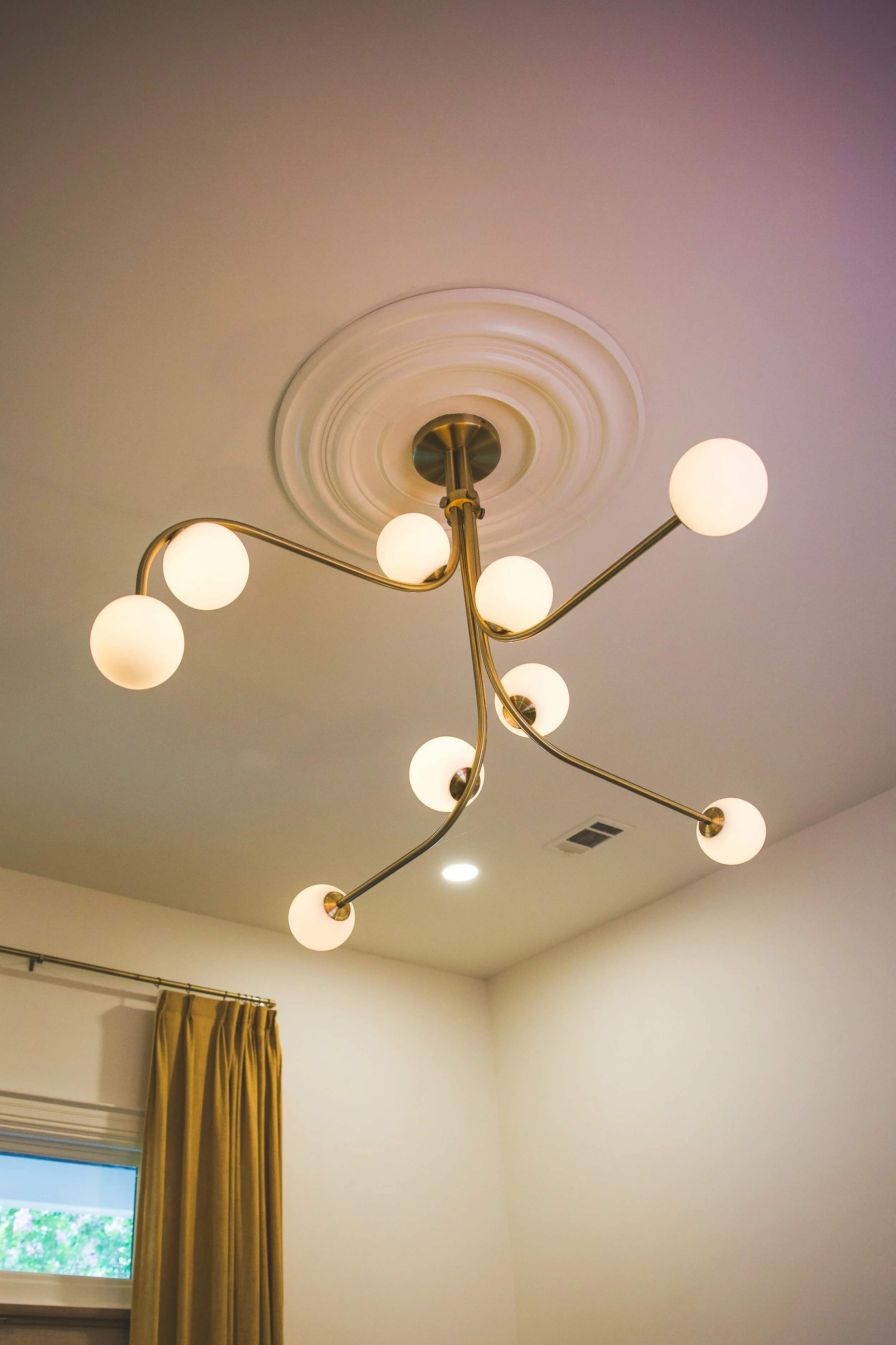 A modern ceiling chandelier with multiple white spherical bulbs on curved brass arms, mounted on a ceiling with decorative ceiling medallion, in a room with beige walls, a window with a mustard-colored curtain, and a small ceiling vent.
