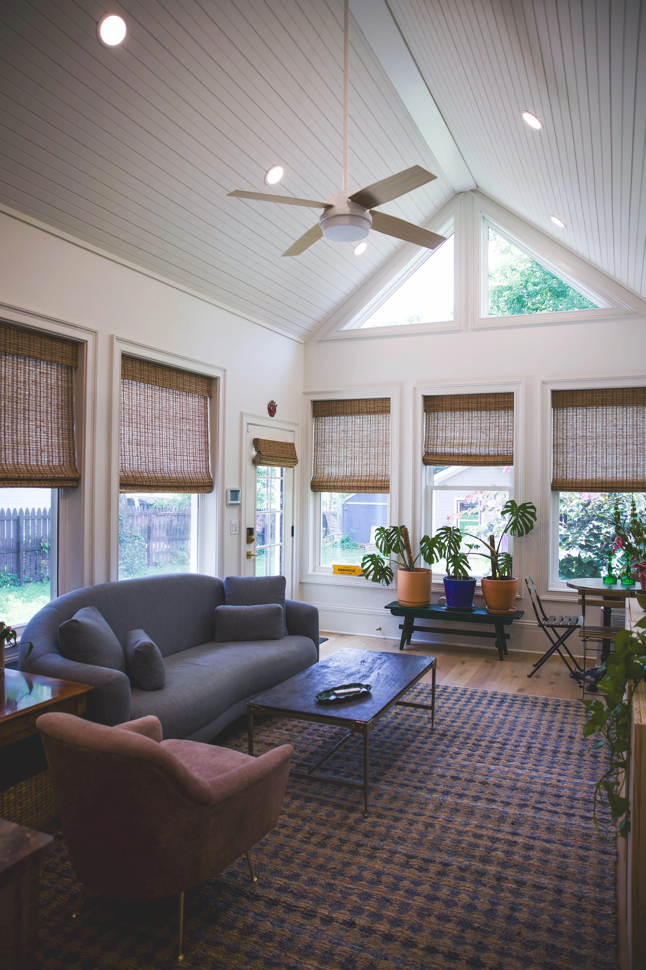 Living room with a vaulted ceiling, ceiling fan, large windows with bamboo shades, and houseplants on a bench.