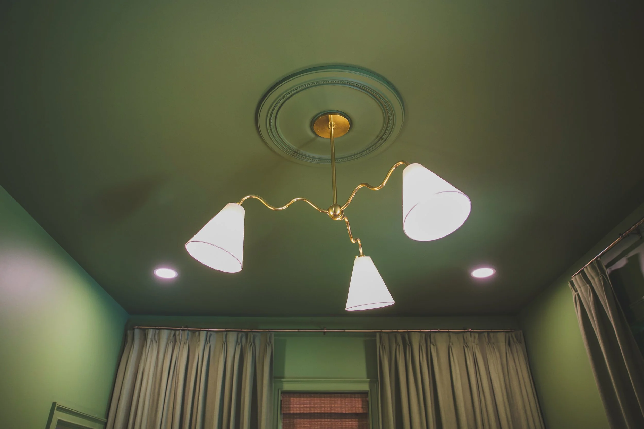 Ceiling with a gold and white chandelier light fixture in a room with green walls, window with curtains, and ceiling lights.