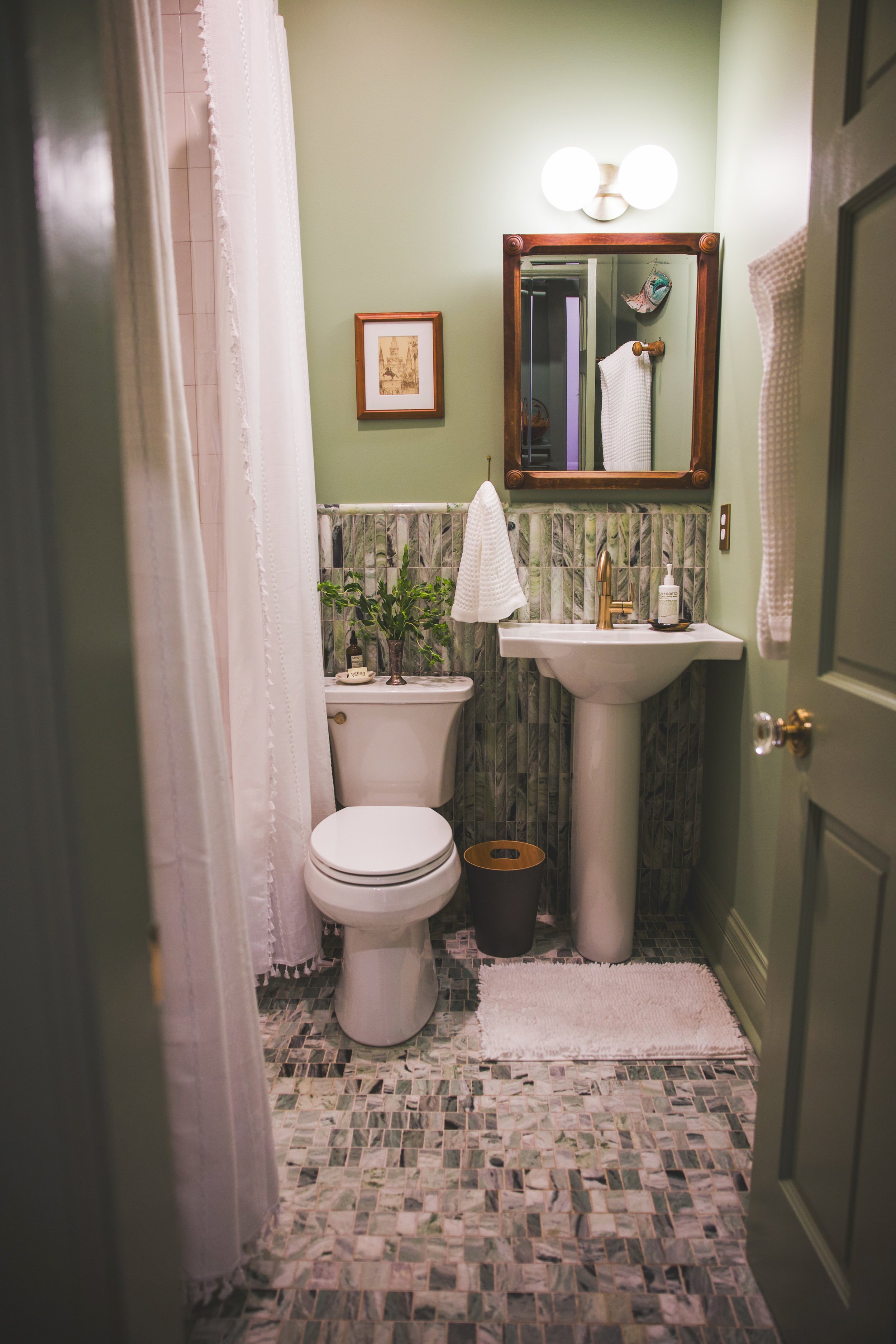 A small bathroom with light green walls and a tile floor. It features a white toilet, a white pedestal sink with a brass faucet, a rectangular wooden-framed mirror, and a framed picture on the wall. A plant and a soap dispenser sit on the lower shelf