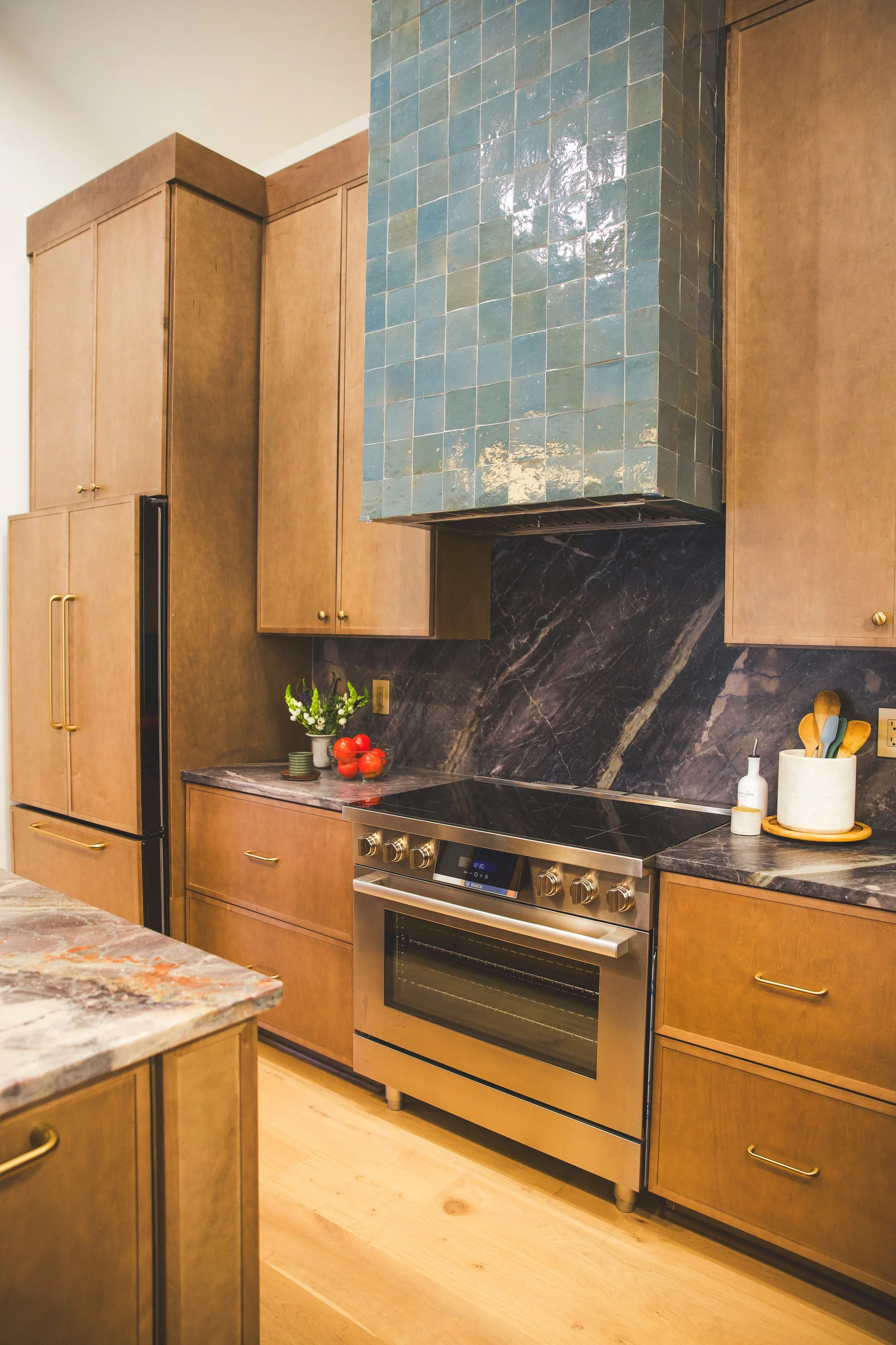Kitchen with wooden cabinets, colorful countertops, a stove, and a light wooden floor. There is a blue tiled chimney above the stove, with a bouquet of flowers, a bowl of tomatoes, and kitchen utensils on the counter.