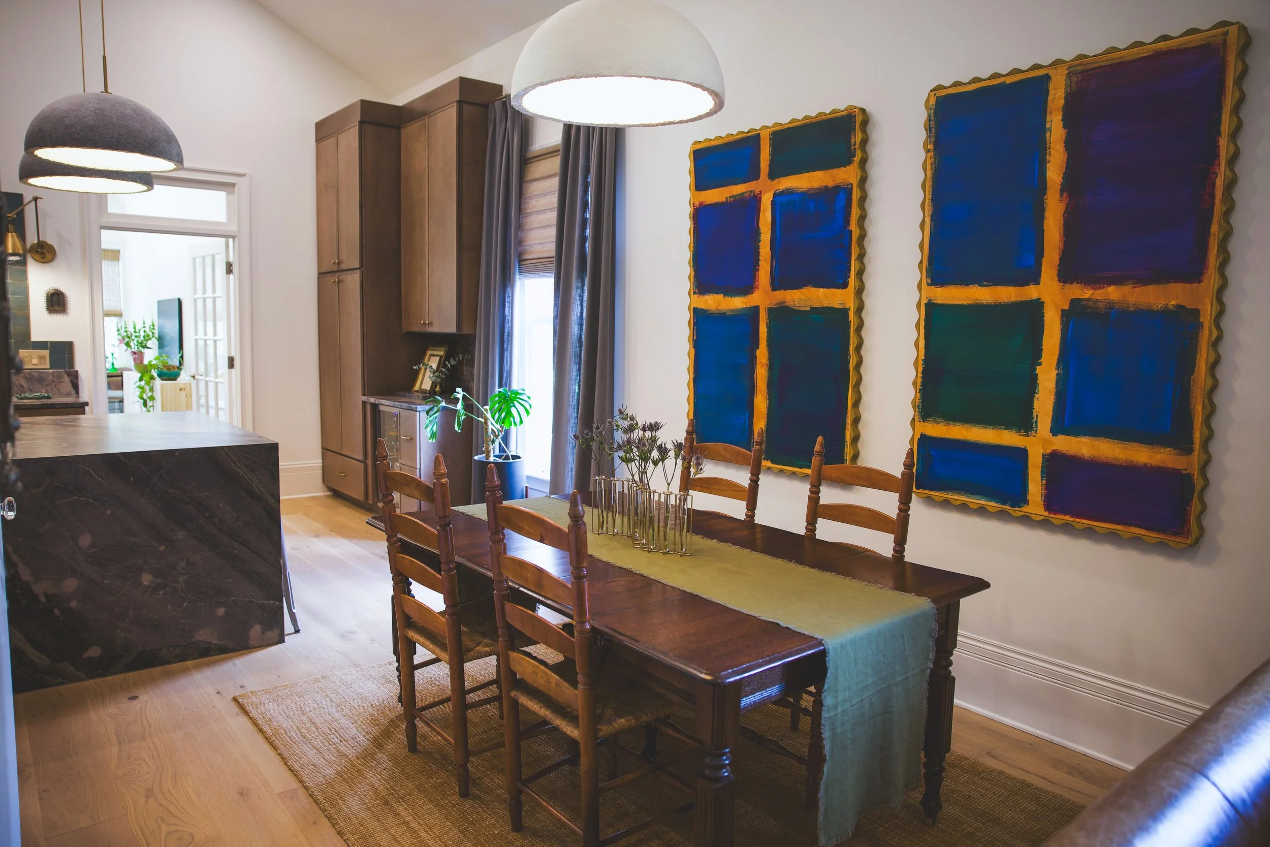 A dining room with a wooden table and six matching chairs, set on a golden rug, with large abstract blue and orange artwork on the wall, and a window with dark curtains.