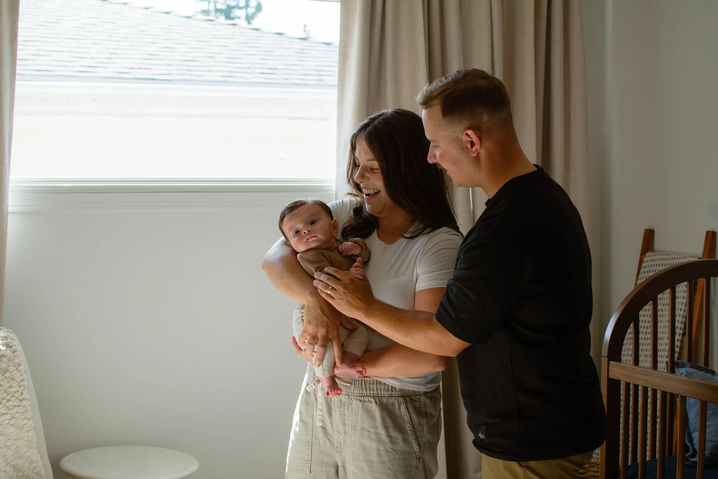 A woman holding an infant with a man standing close by, indoors near a window with curtains.