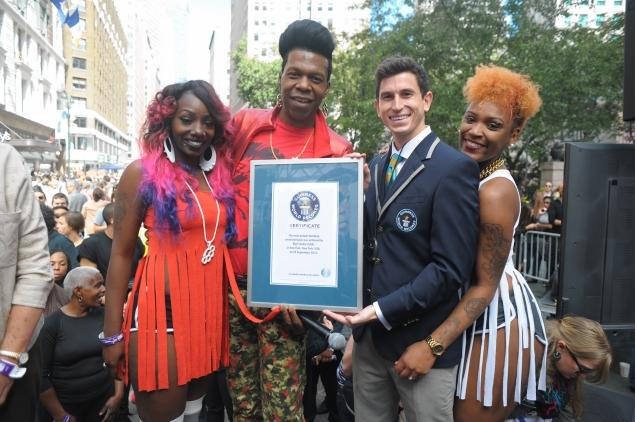 Four diverse people smiling and holding a framed certificate outdoors in an urban setting, with a crowd and buildings in the background.