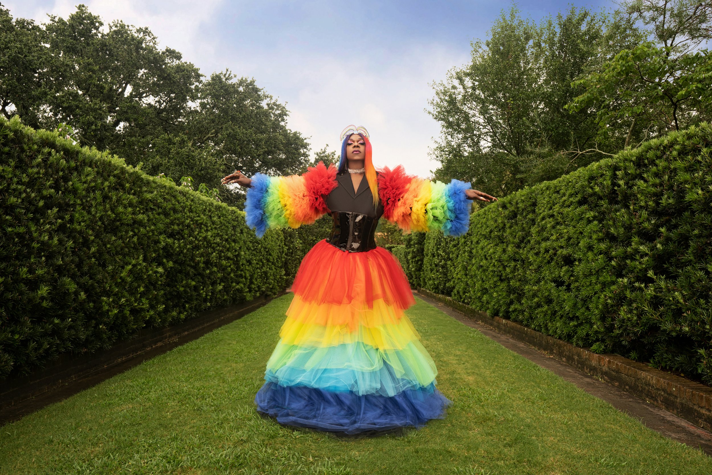 Person standing in a garden wearing a multicolored rainbow dress with puffy sleeves and a black corset, with arms outstretched and a rainbow-colored hairstyle.
