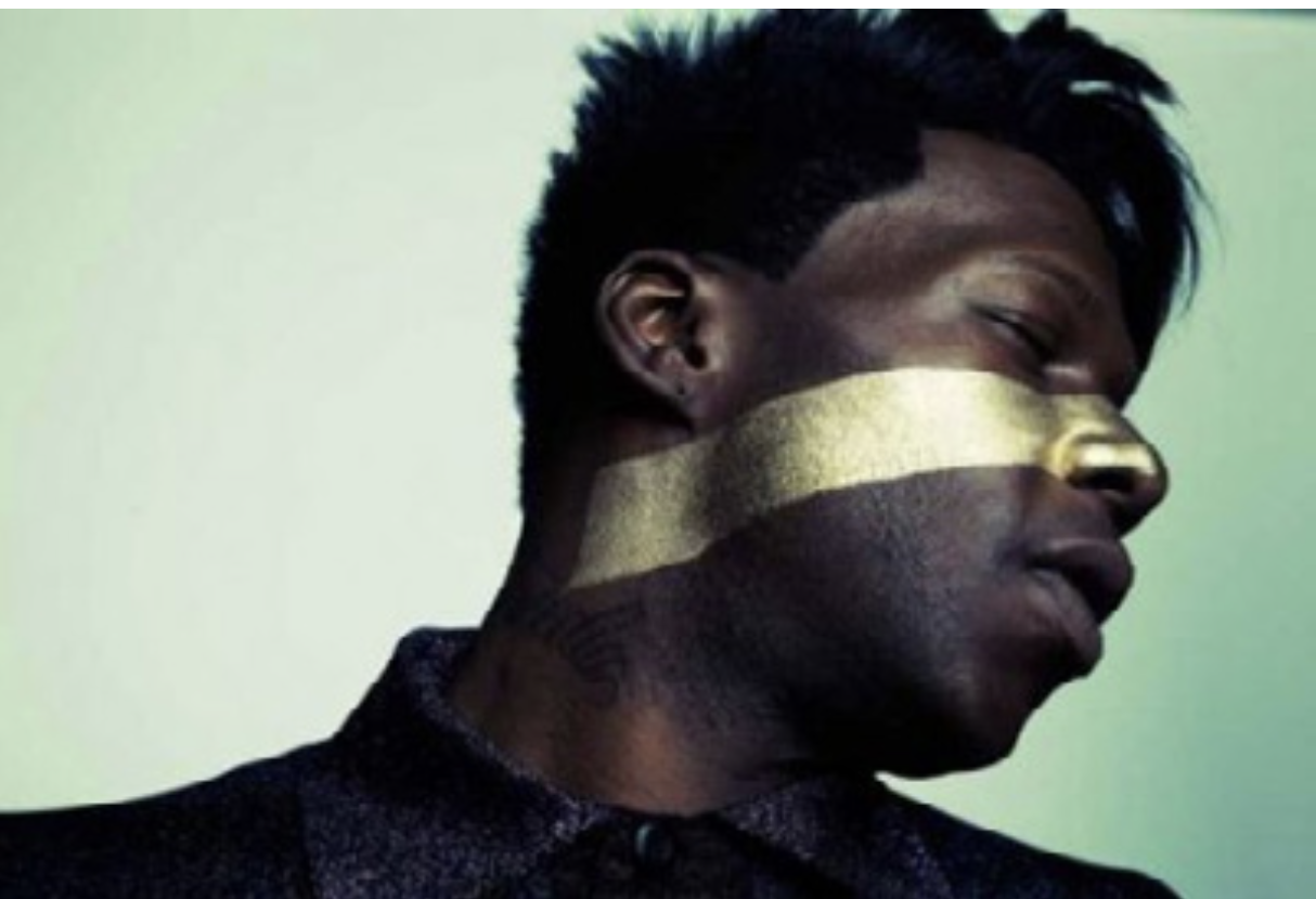 A person with dark skin and short, spiky black hair is facing sideways with their eyes closed. They have a wide metallic gold stripe painted across their face and neck, and wear a dark, textured shirt.