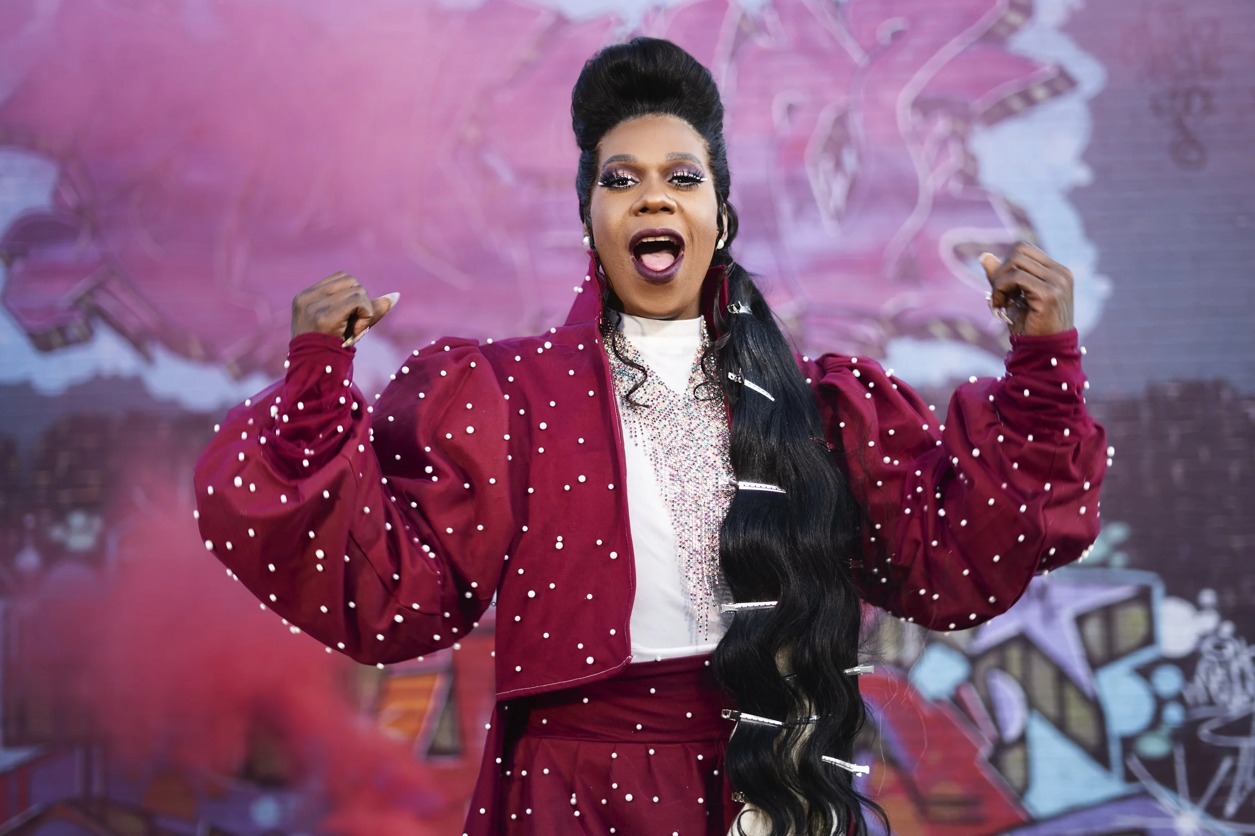 A woman wearing a red jacket with pearl embellishments, raising her flexed arms confidently, in front of a pink and blue graffiti background.