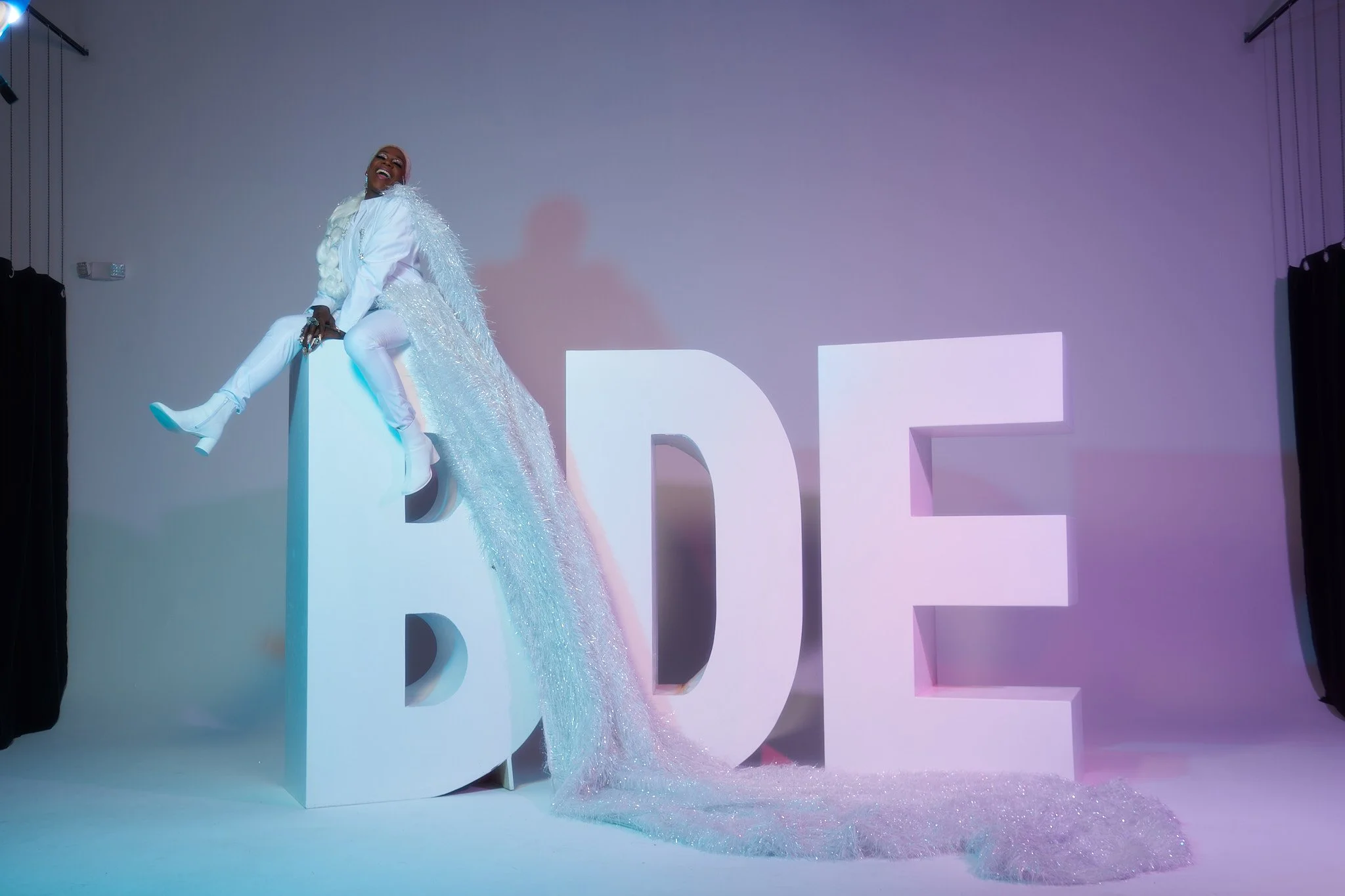 Person dressed in white sitting on large white letters spelling 'LOVE' with a sparkly cape, in a studio with purple lighting.