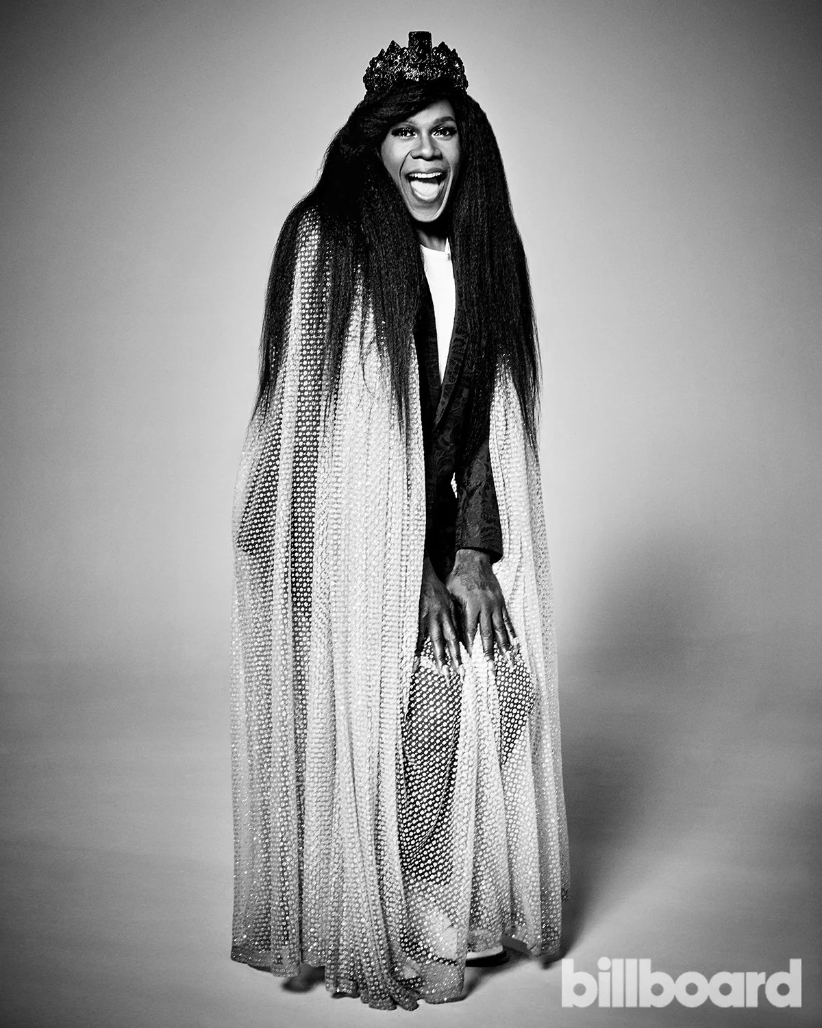 Black and white photo of a woman wearing a crown, a long flowing cape, and formal attire, smiling with her hands on her thighs against a plain background.