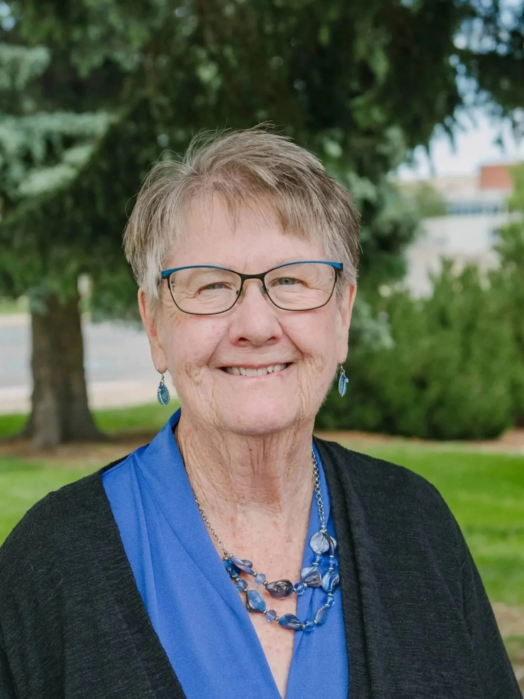 A smiling elderly woman with short gray hair and glasses standing outdoors with trees in the background. She is wearing a blue blouse, a black cardigan, and jewelry including a necklace and earrings.