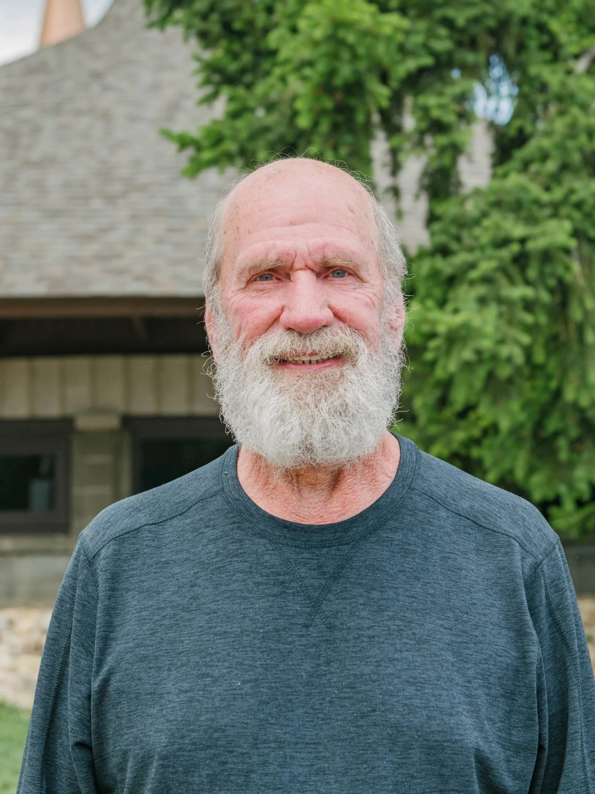 Close-up of an elderly man with a full white beard, wearing a dark gray shirt, standing outdoors in front of green trees and a building with a sloped roof.