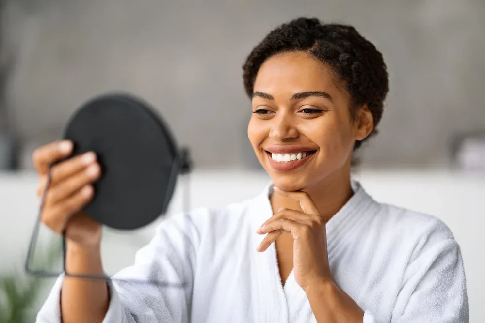Woman smiling while examining her new dental veneers in a handheld mirror at Boulder clinic.