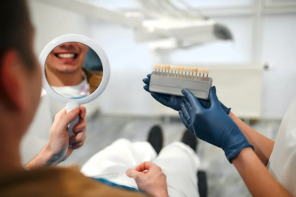  Dentist showing patient shade guide with mirror reflection to compare veneer options during consultation