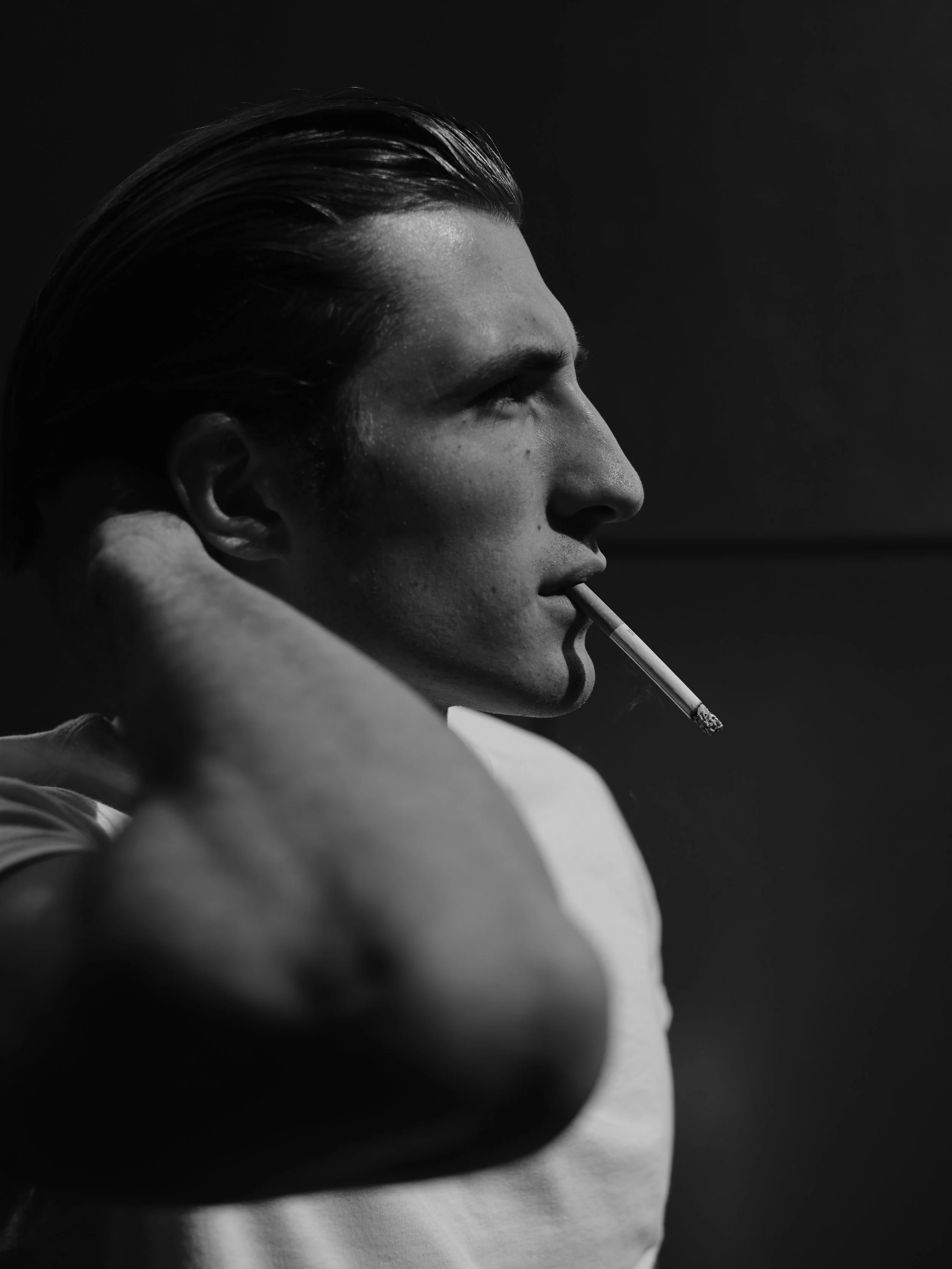 Black and white photo of a man with slicked-back hair smoking a cigarette, facing to the right, with a contemplative expression.