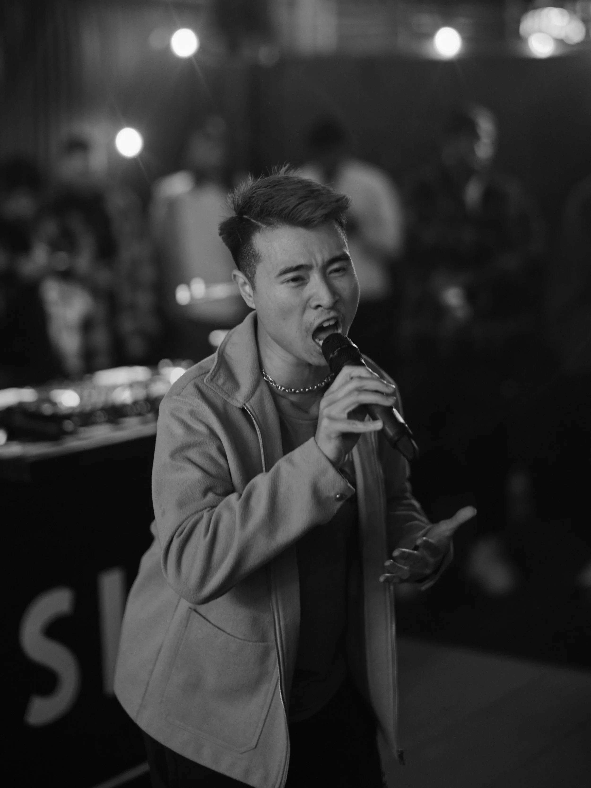A young man singing into a microphone at a lively indoor gathering, with people and DJ equipment in the background.