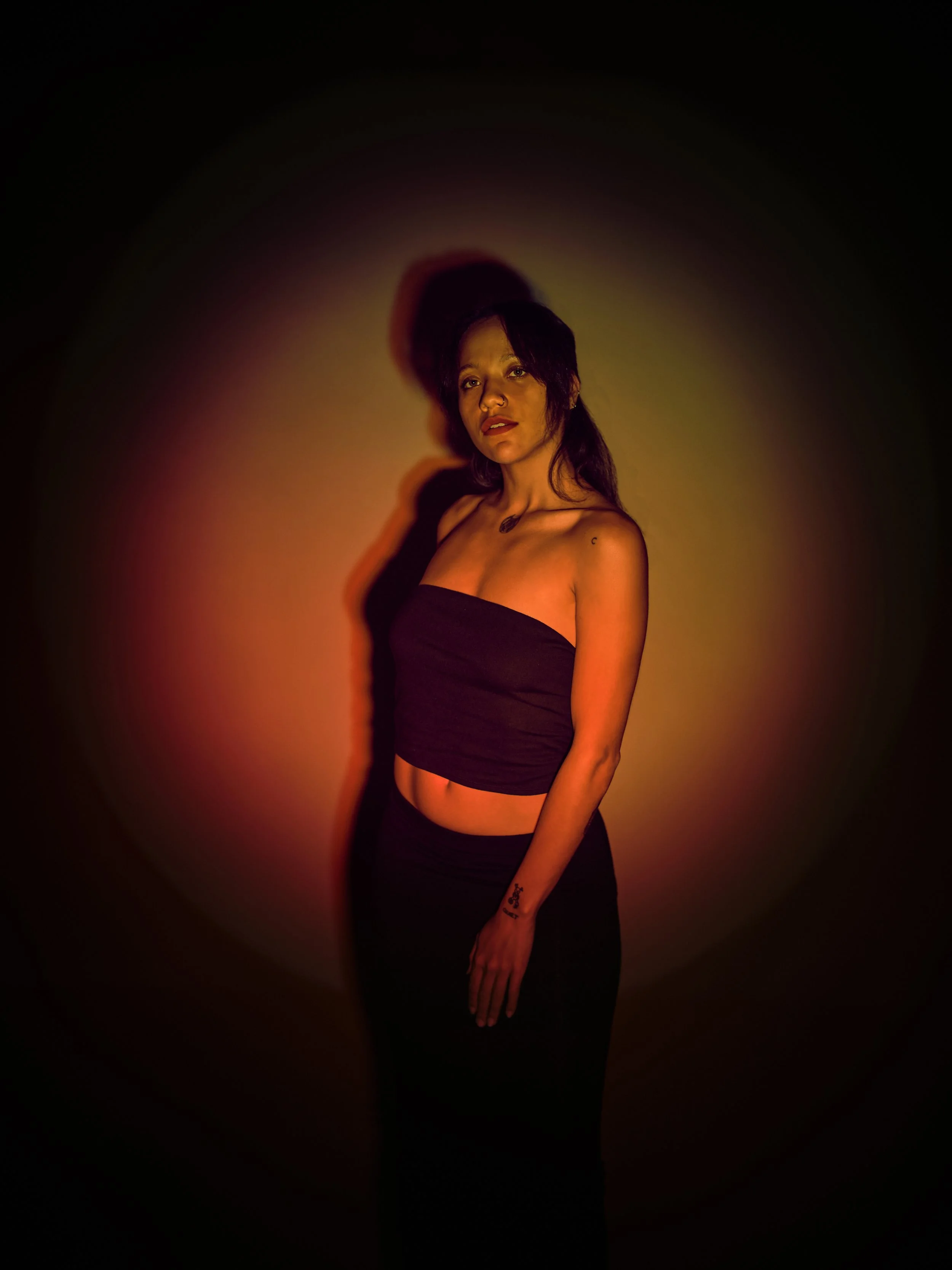 A woman with dark hair wearing a strapless black top and black pants standing against a multicolored light background, casting a shadow on the wall.