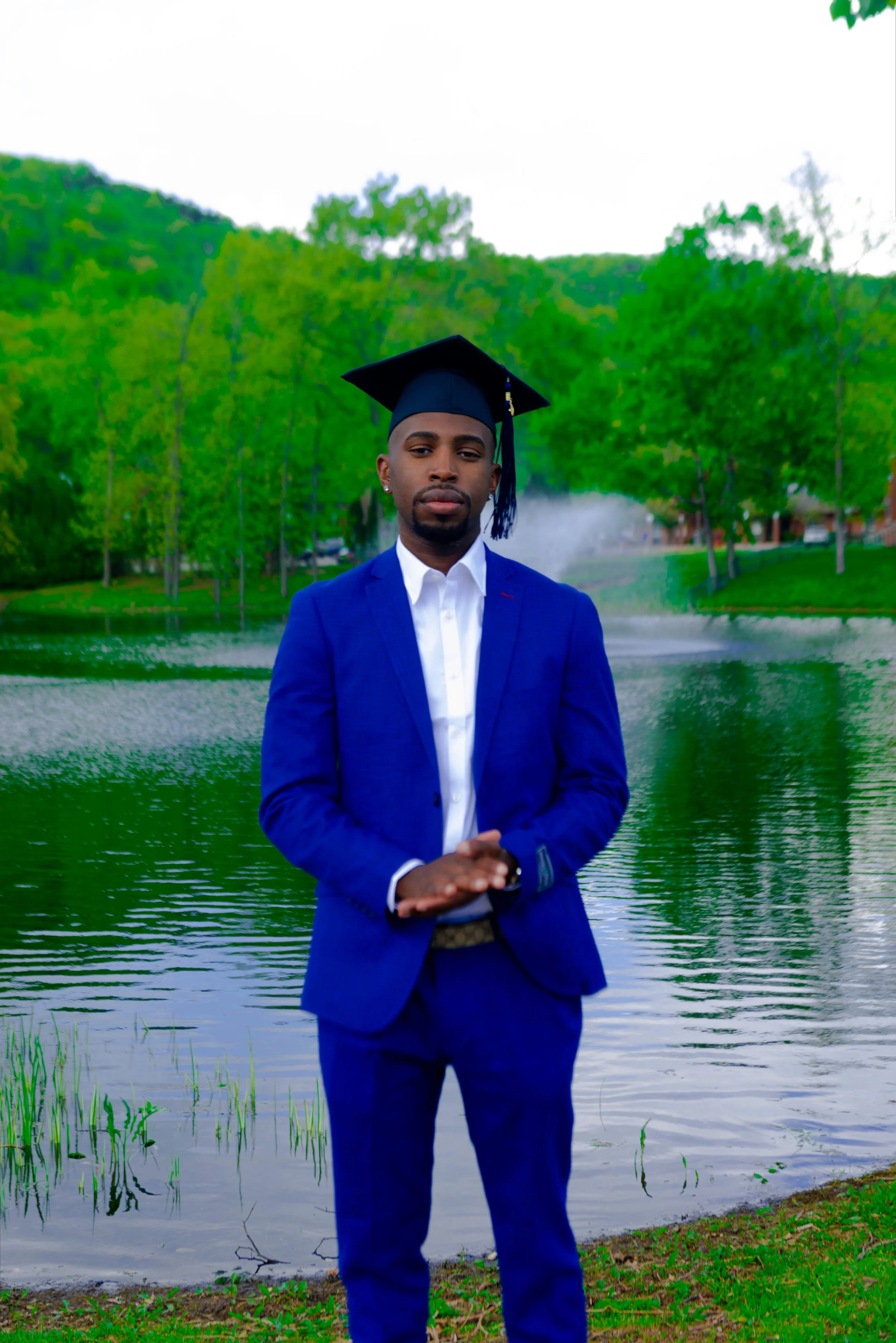 Young man in a blue suit and white shirt wearing a graduation cap, standing near a lake with green trees and a fountain in the background.
