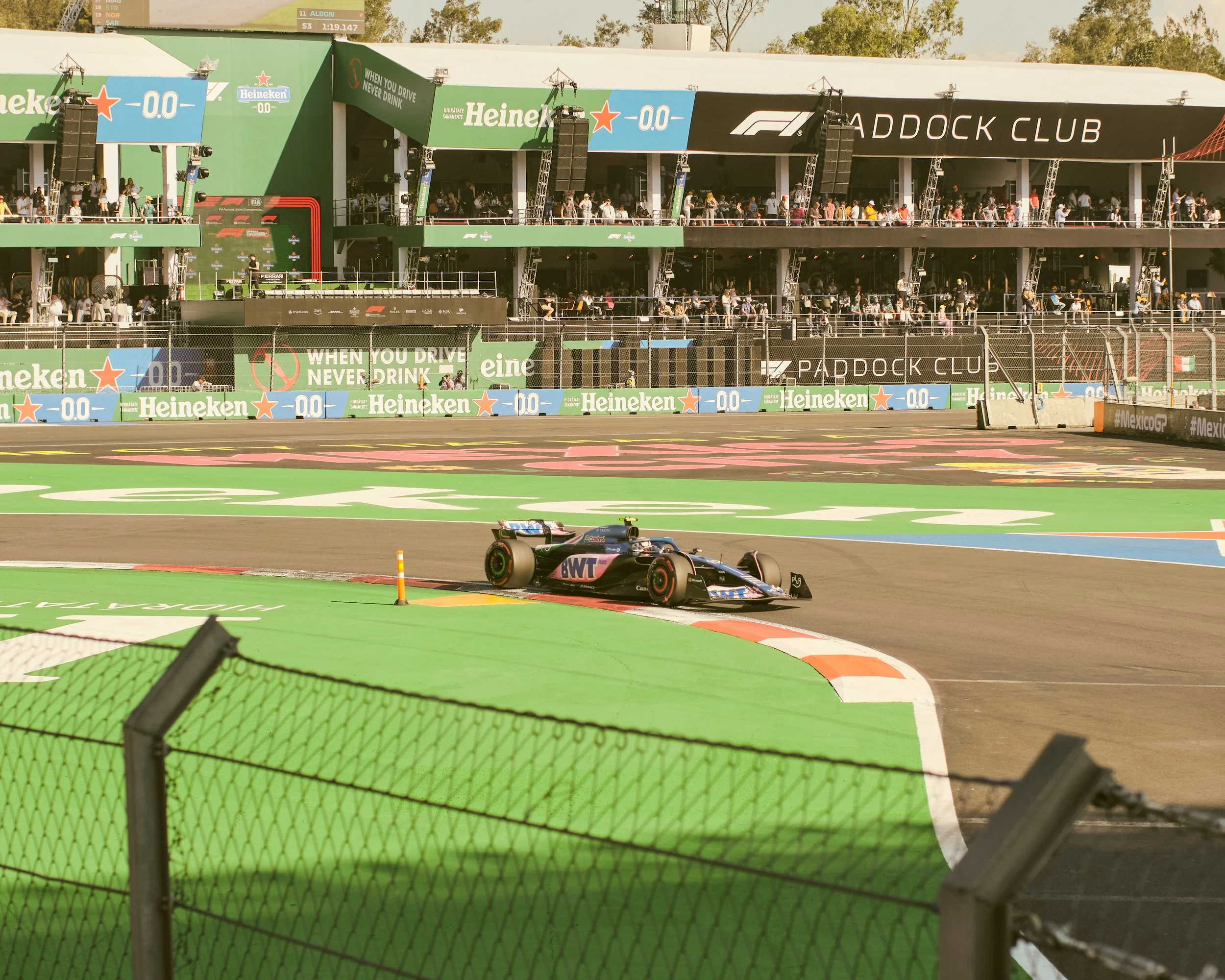 A Formula 1 race car rounding a corner at the Autódromo Hermanos Rodríguez during a Grand Prix, with the Paddock Club and spectators in the background and advertising banners along the track.
