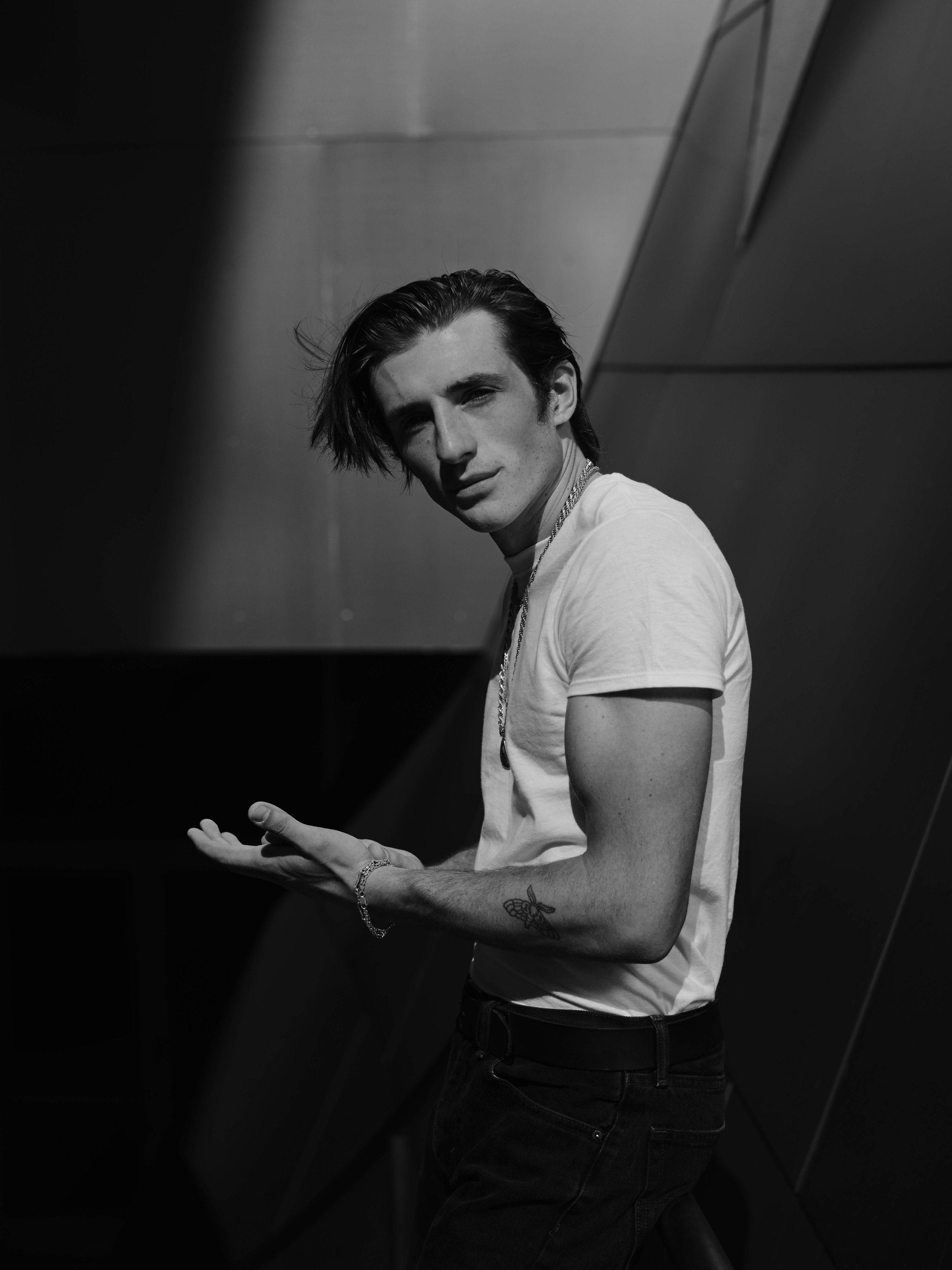 A young man with dark, tousled hair, wearing a white t-shirt and dark jeans, is posing against a metallic background with geometric panels, looking at the camera in black and white.