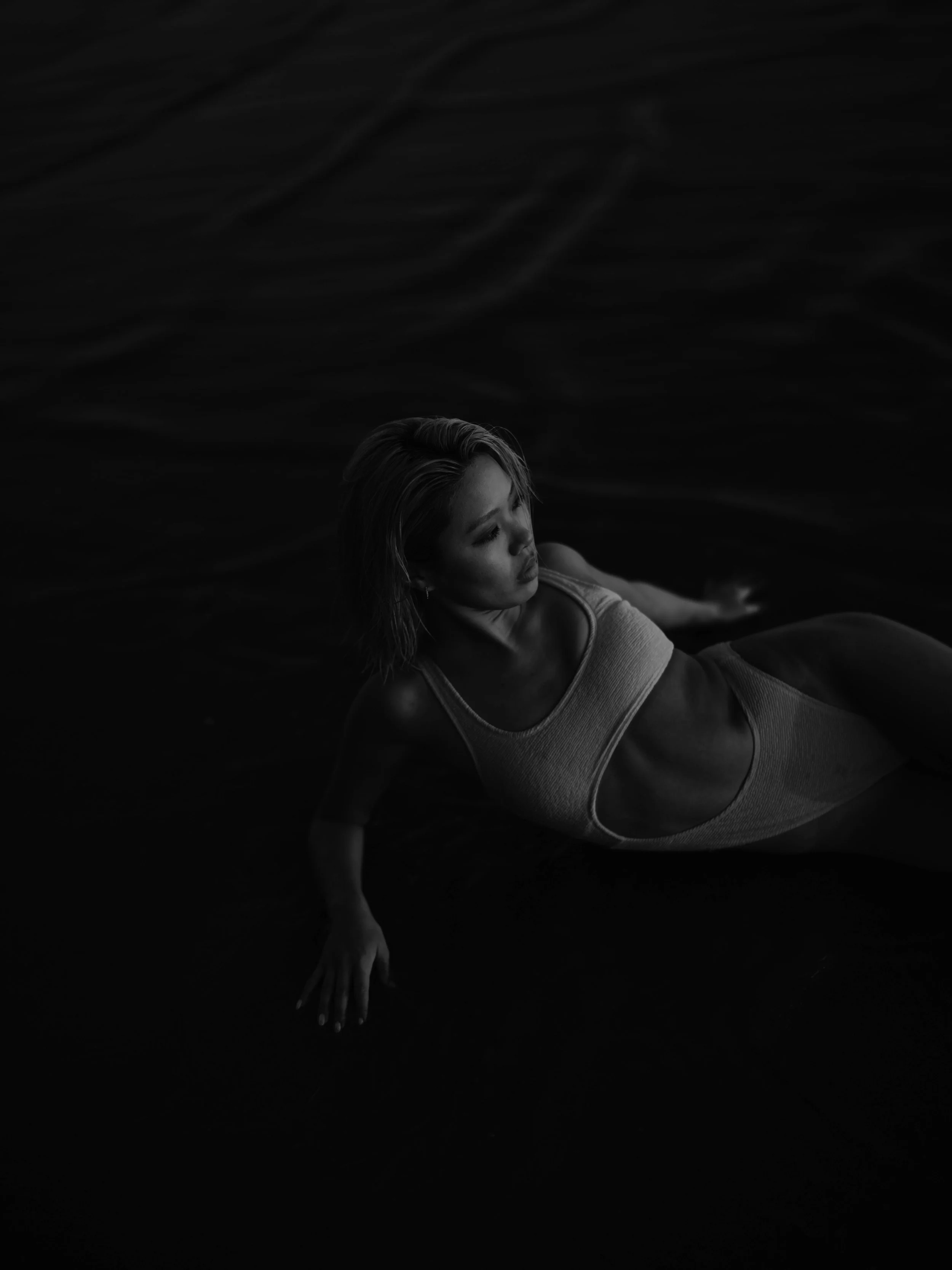 A woman with short hair reclining on a dark surface, wearing a sleeveless top with a circular cutout in the midsection, looking contemplative in a monochromatic photograph.