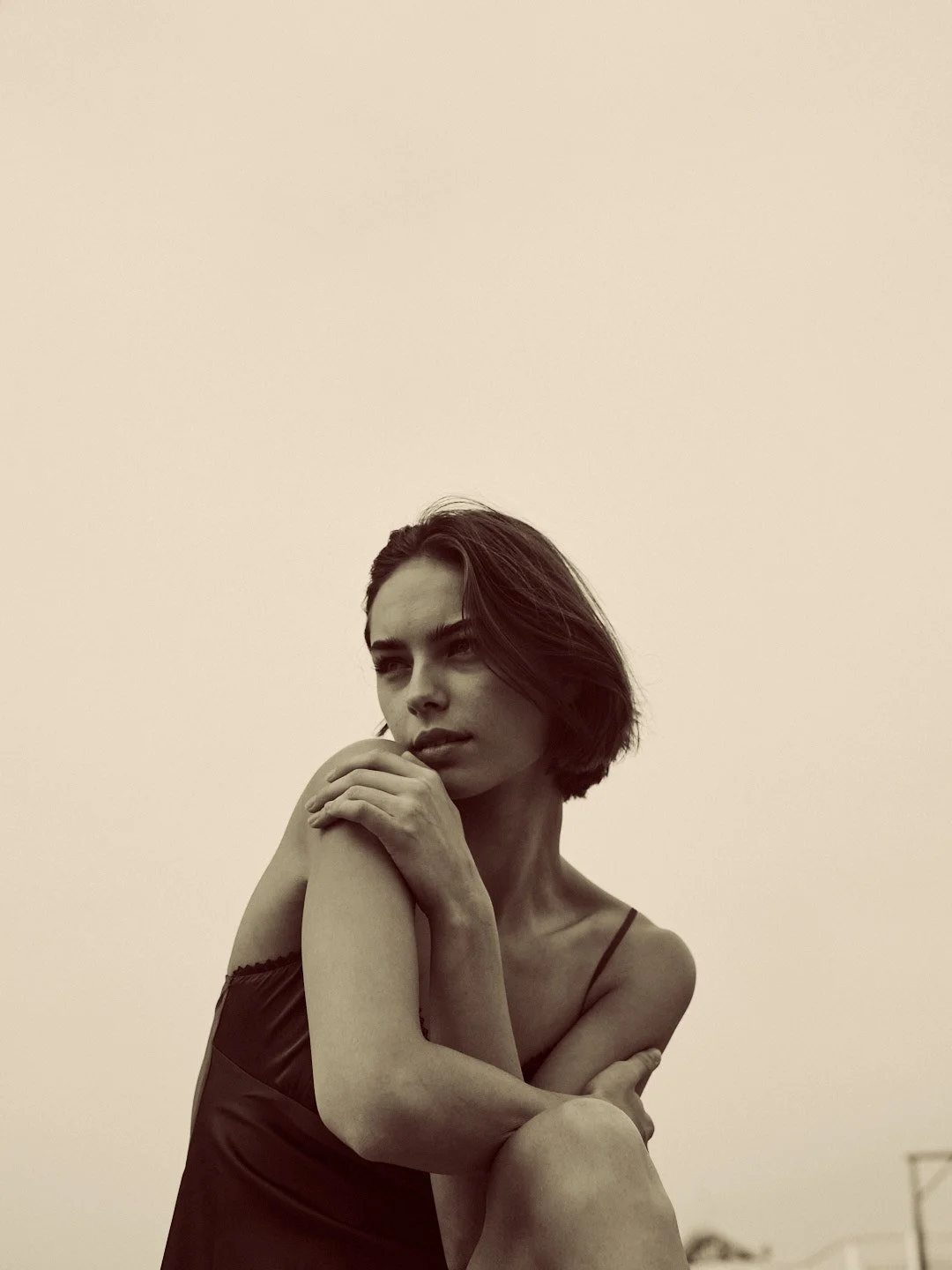 A woman with short dark hair and a serious expression, wearing a dark spaghetti strap top, sitting with her arms crossed, in a minimalist setting with a plain background.