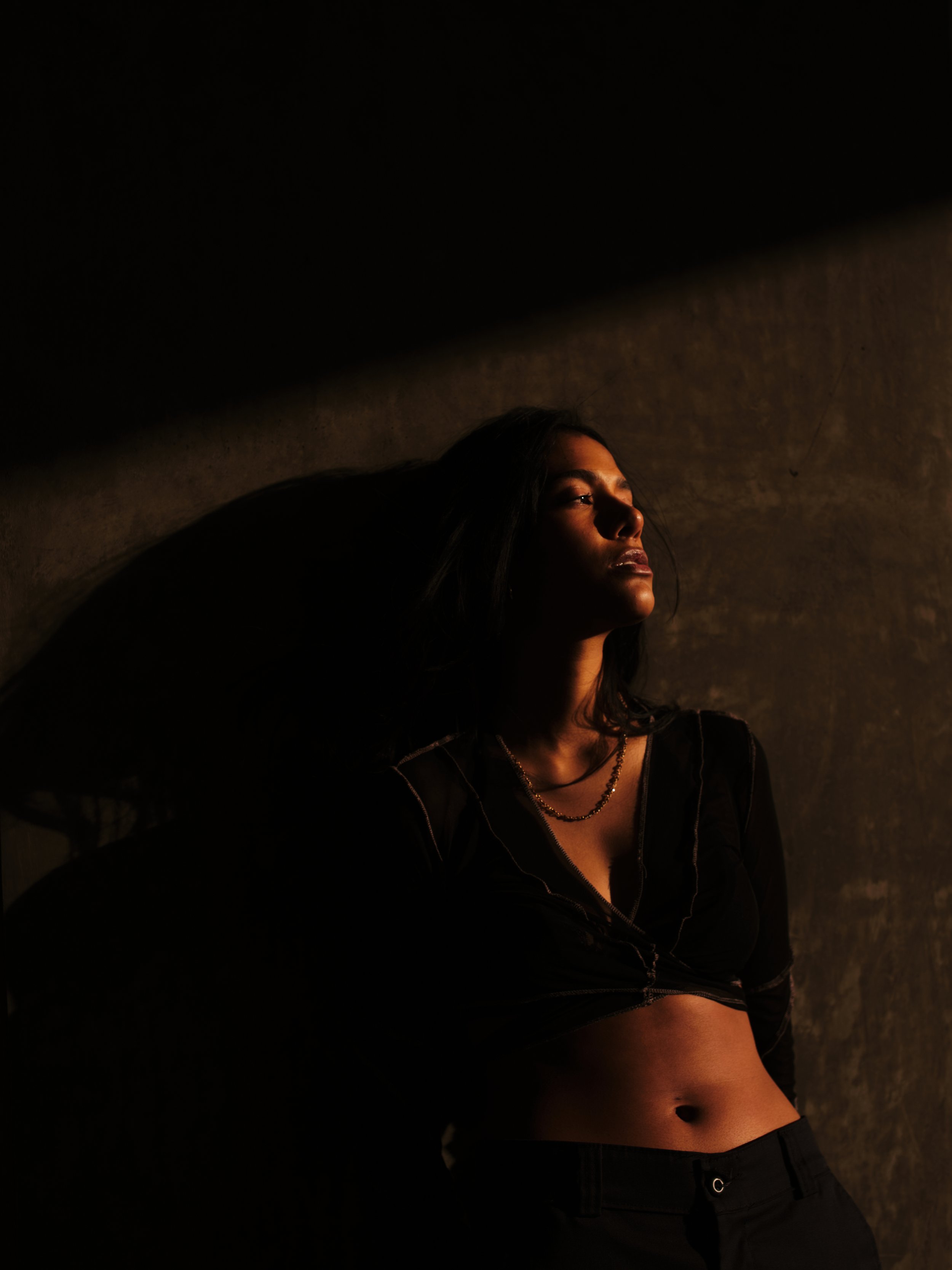 A woman with dark hair and dark clothing leaning against a wall in low lighting, with dramatic shadows.
