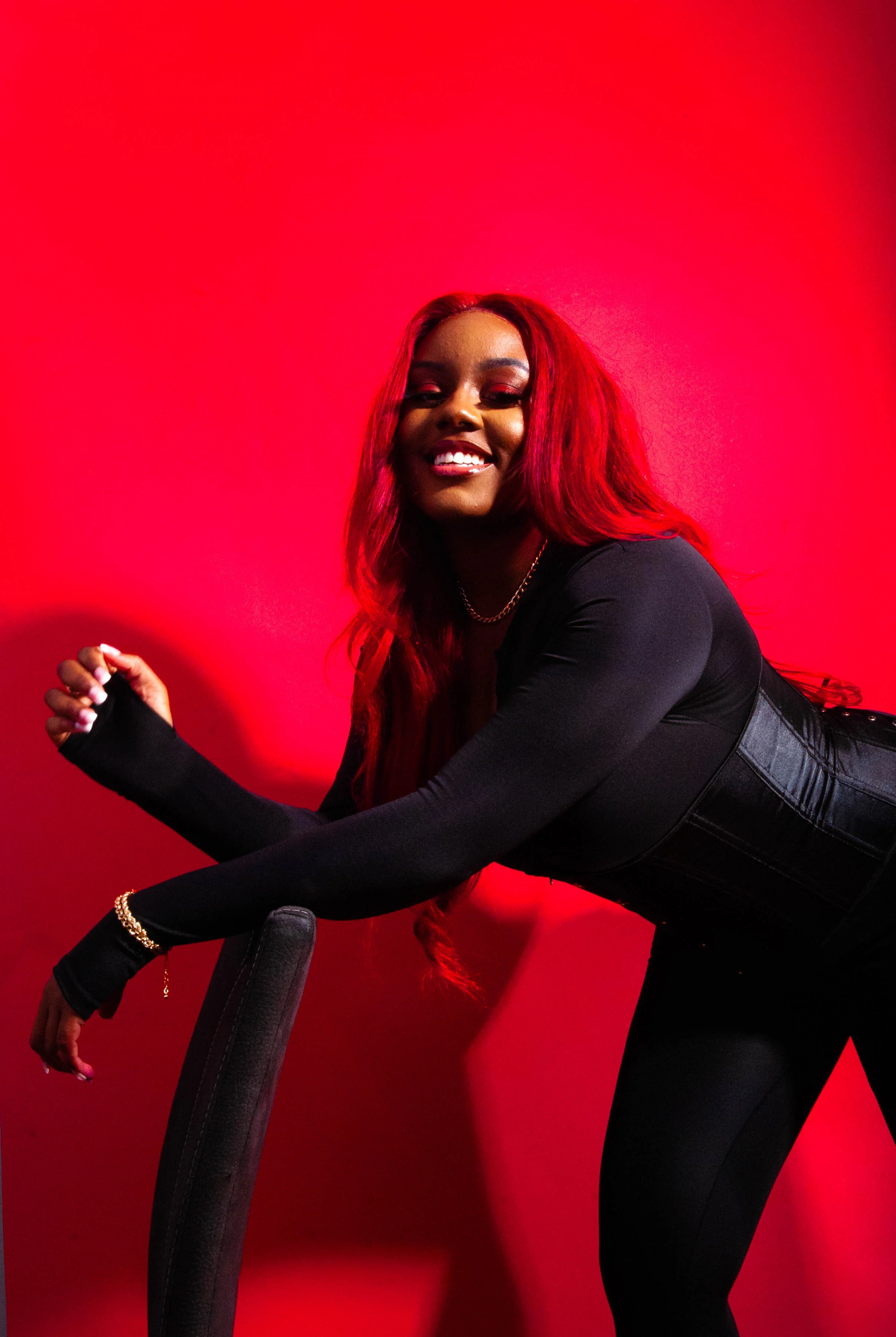 A smiling woman with long red hair, wearing a black outfit, posing against a bright red background.