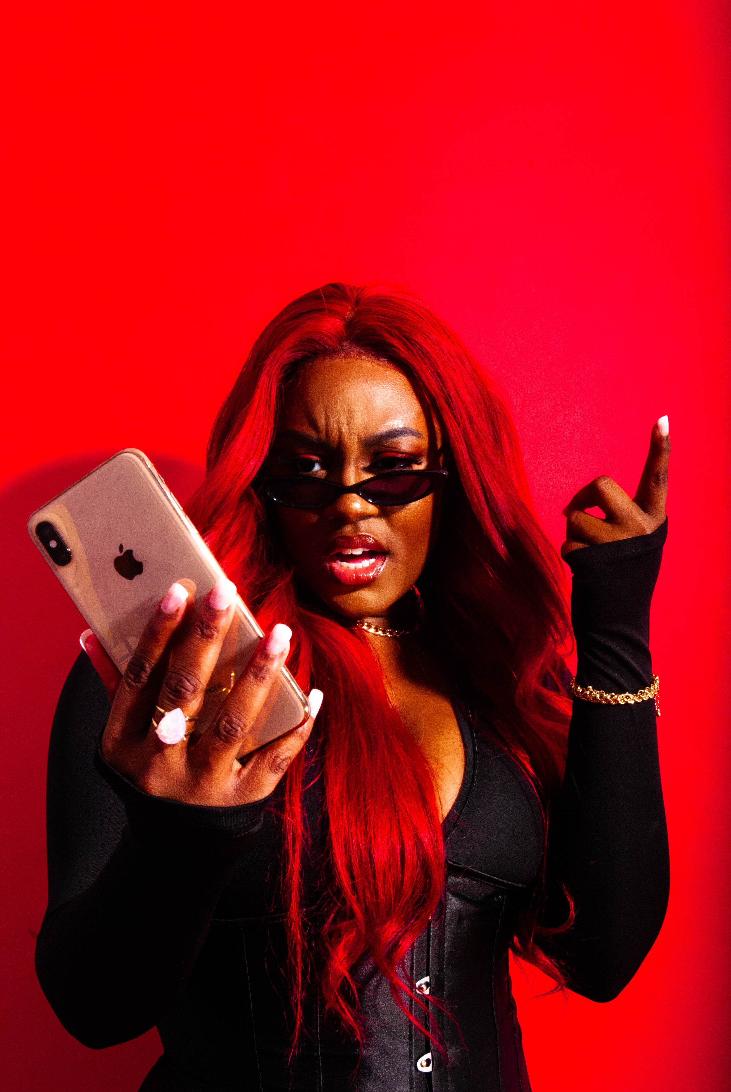 Young woman with red hair wearing black sunglasses, holding a smartphone, and pointing her finger with a confident expression against a red background.