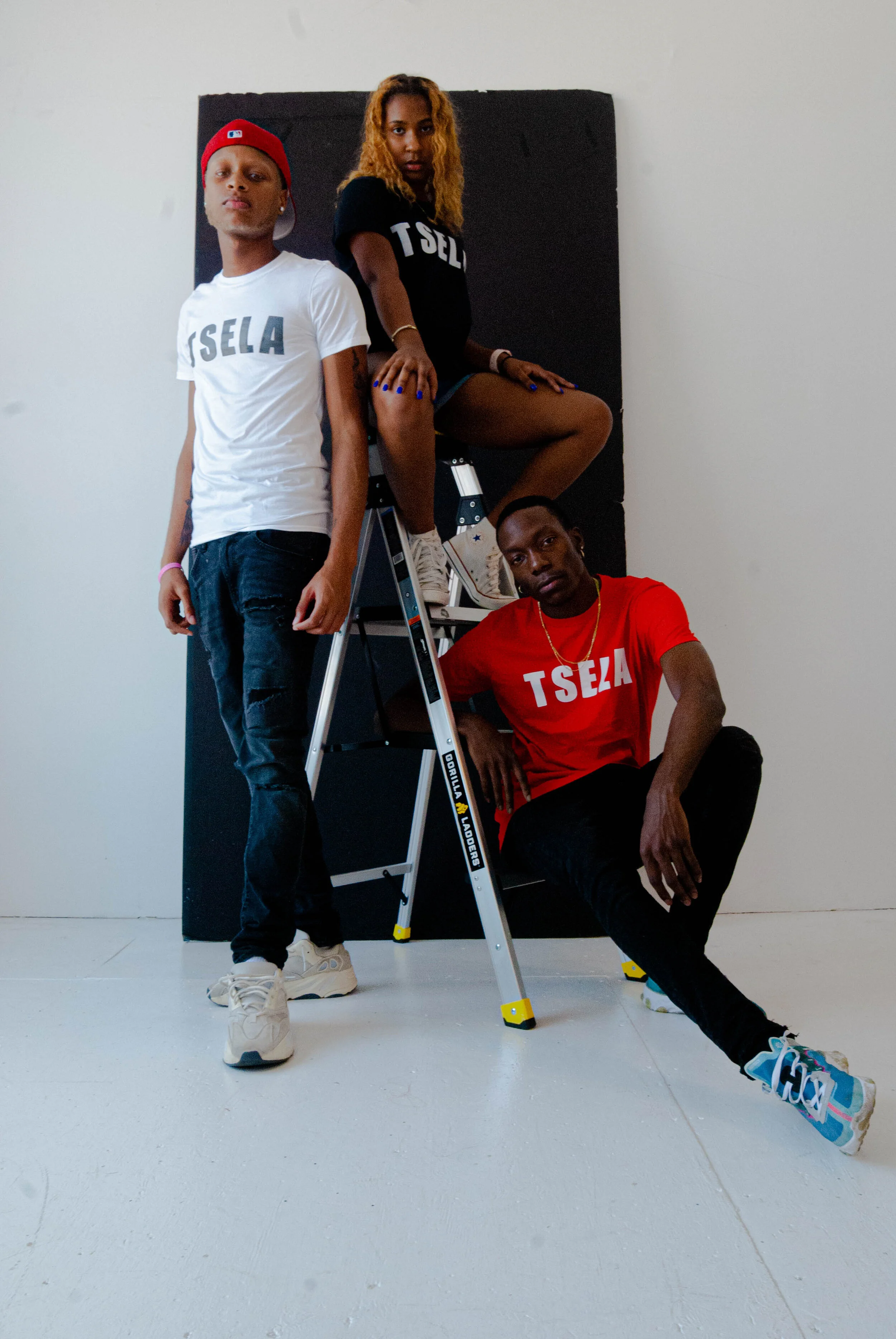 Group of three young adults posing indoors. One person is standing, another sitting on a ladder, and the third sitting on the floor. They are wearing casual streetwear, including T-shirts with 