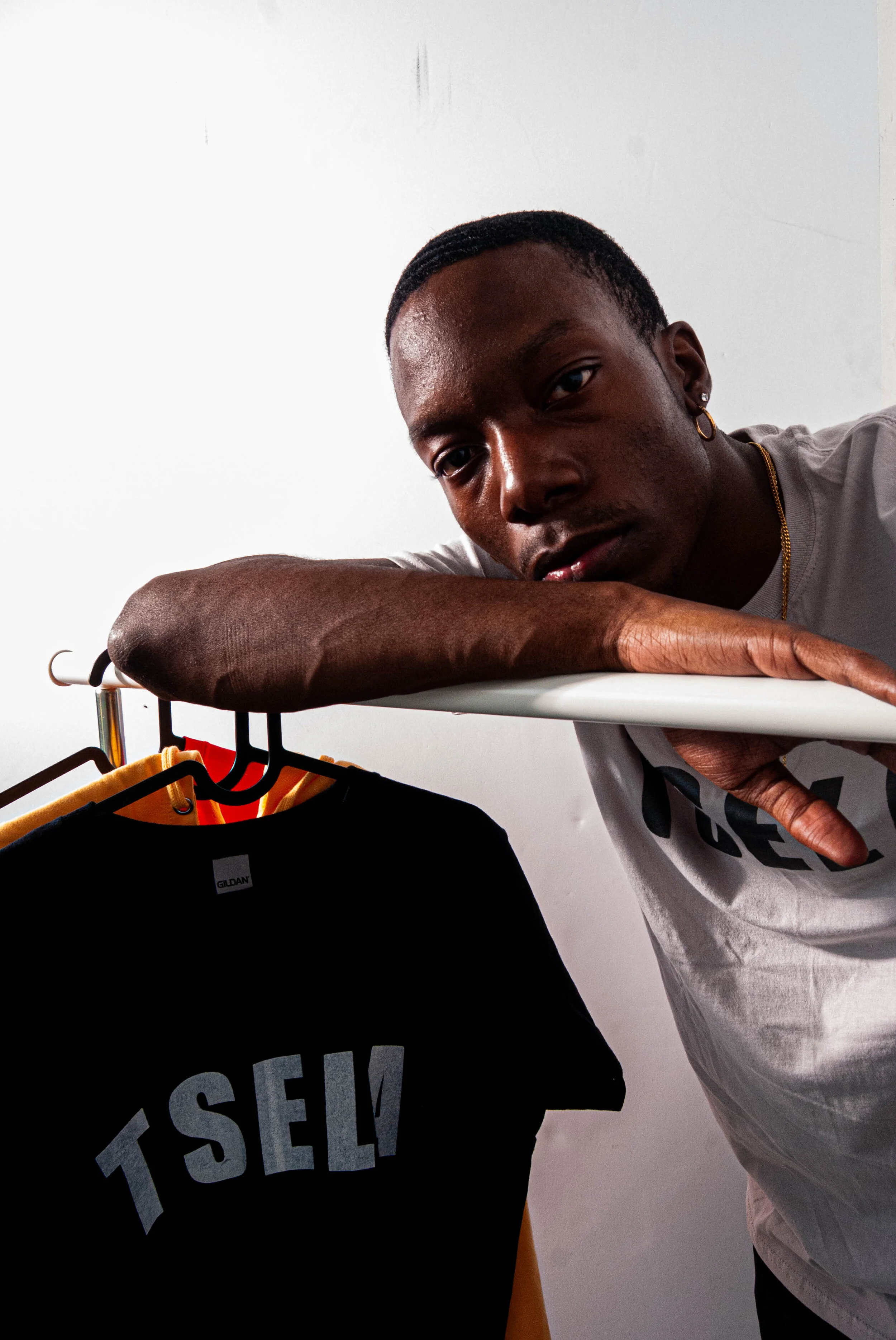 A young man leaning on a white shelf with a clothing rack below him. The rack has black and yellow shirts, with one shirt displaying the words "I SELF!" in large gray letters.