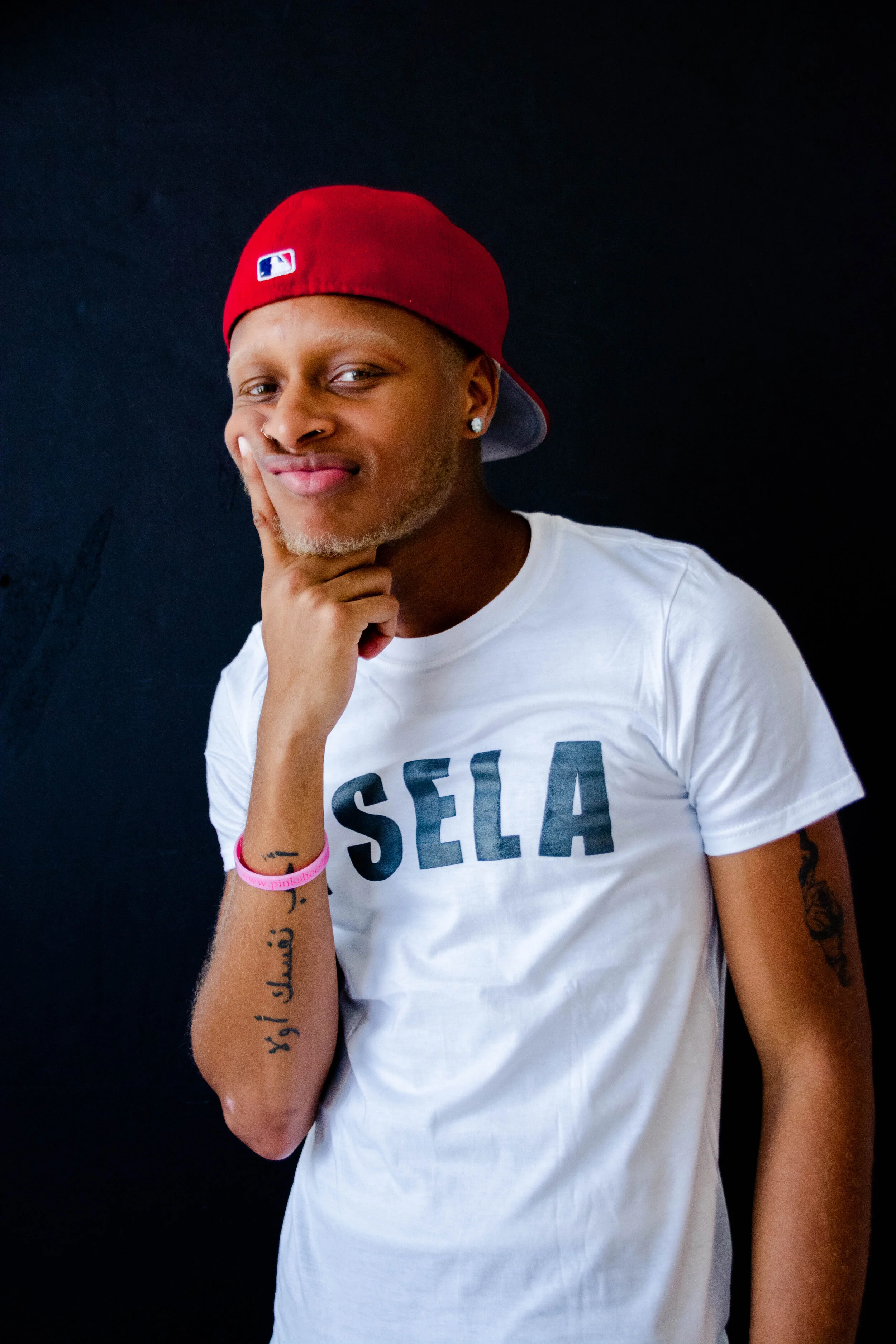 A man wearing a red baseball cap backwards, a white Sela T-shirt, and showing tattoos on his left arm, standing against a dark background.