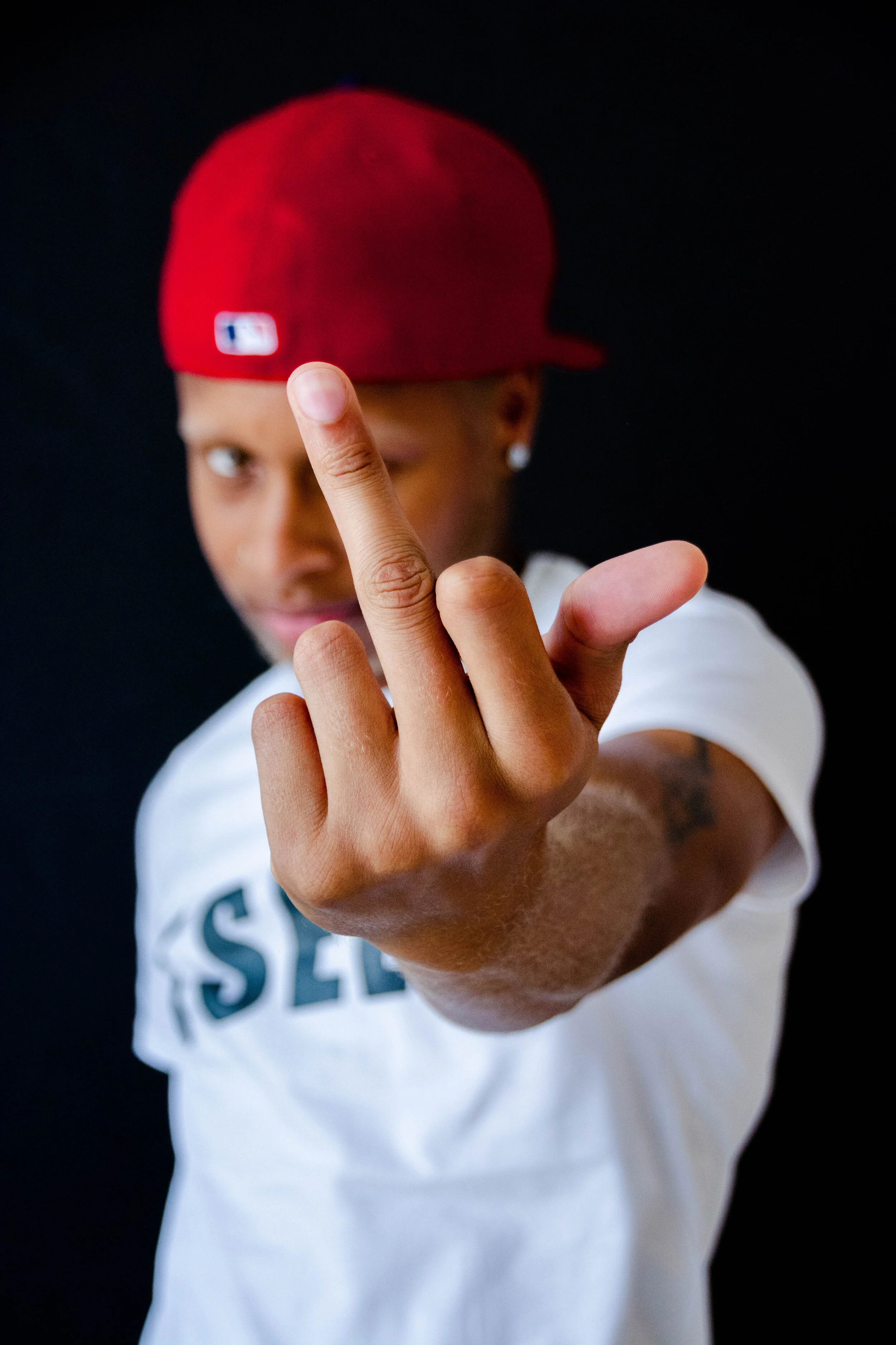 A young man wearing a red baseball cap and a white shirt, giving the middle finger gesture towards the camera with a black background.