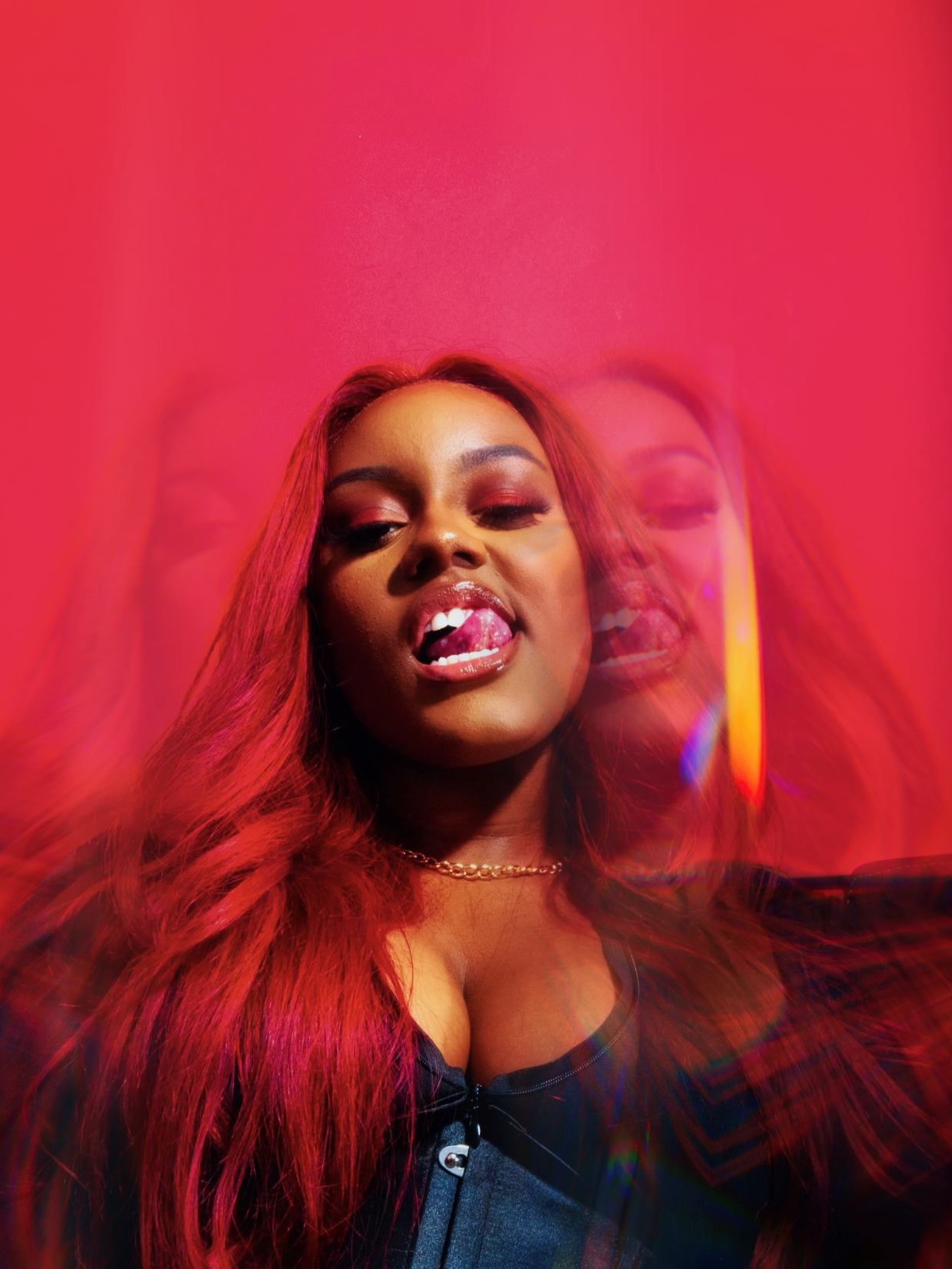 A woman with long red hair making a playful expression with her tongue out, against a pink background.